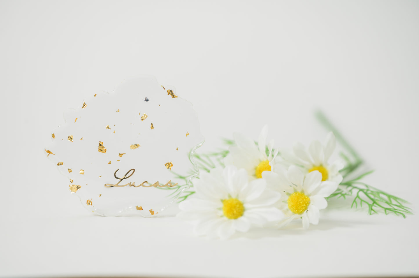 A clear, irregular-shaped resin Coaster with gold foil accents, featuring the name 'Lucas' in elegant calligraphy, displayed alongside white daisies for a fresh, sophisticated look.
