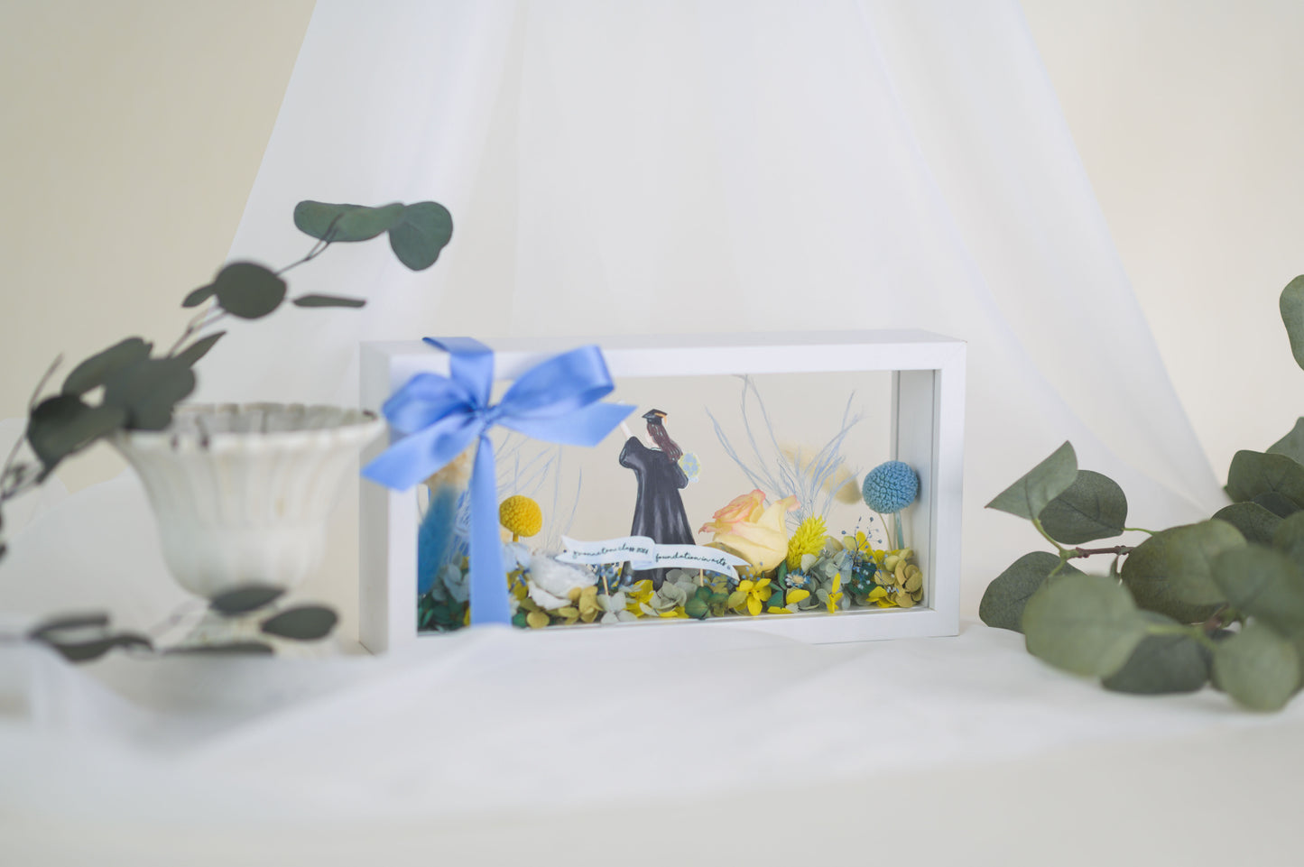 Graduation-themed preserved flower frame featuring a graduate figure holding a diploma, surrounded by vibrant yellow, blue, and white blooms.