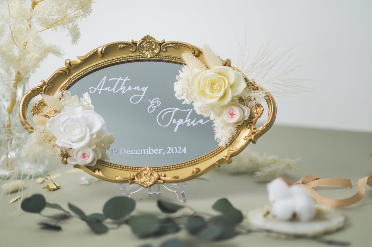 Ornate gold-framed wedding mirror featuring preserved white flowers, with customizable names Anthony & Sophia and wedding date, beautifully displayed on a reception table with floral accents.