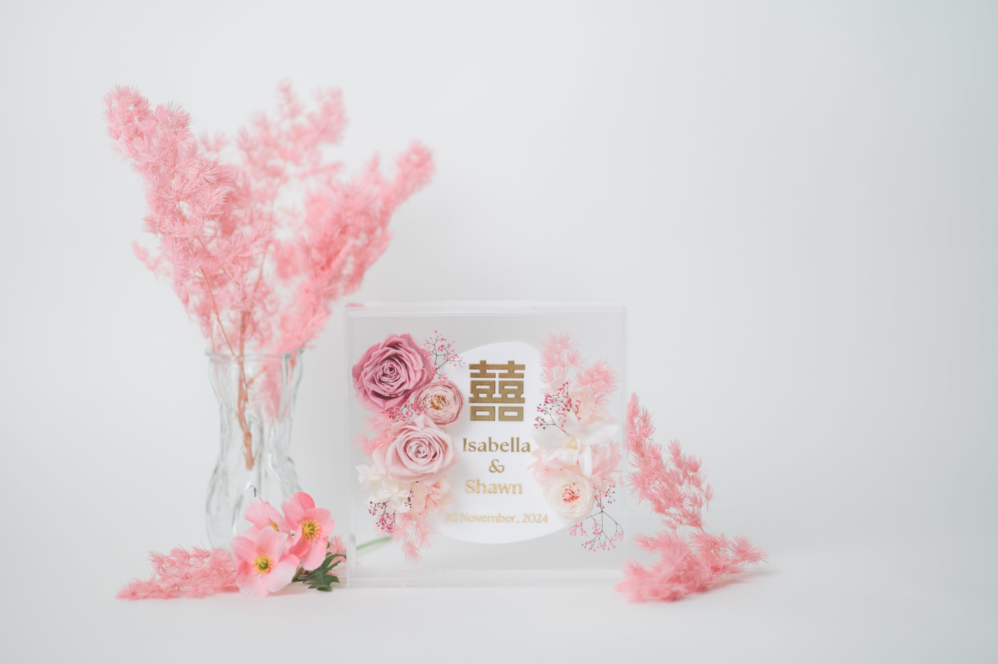 Personalized wedding frame with preserved pink flowers, featuring custom names Isabella & Shawn and a wedding date, with elegant floral design in a clear acrylic display.