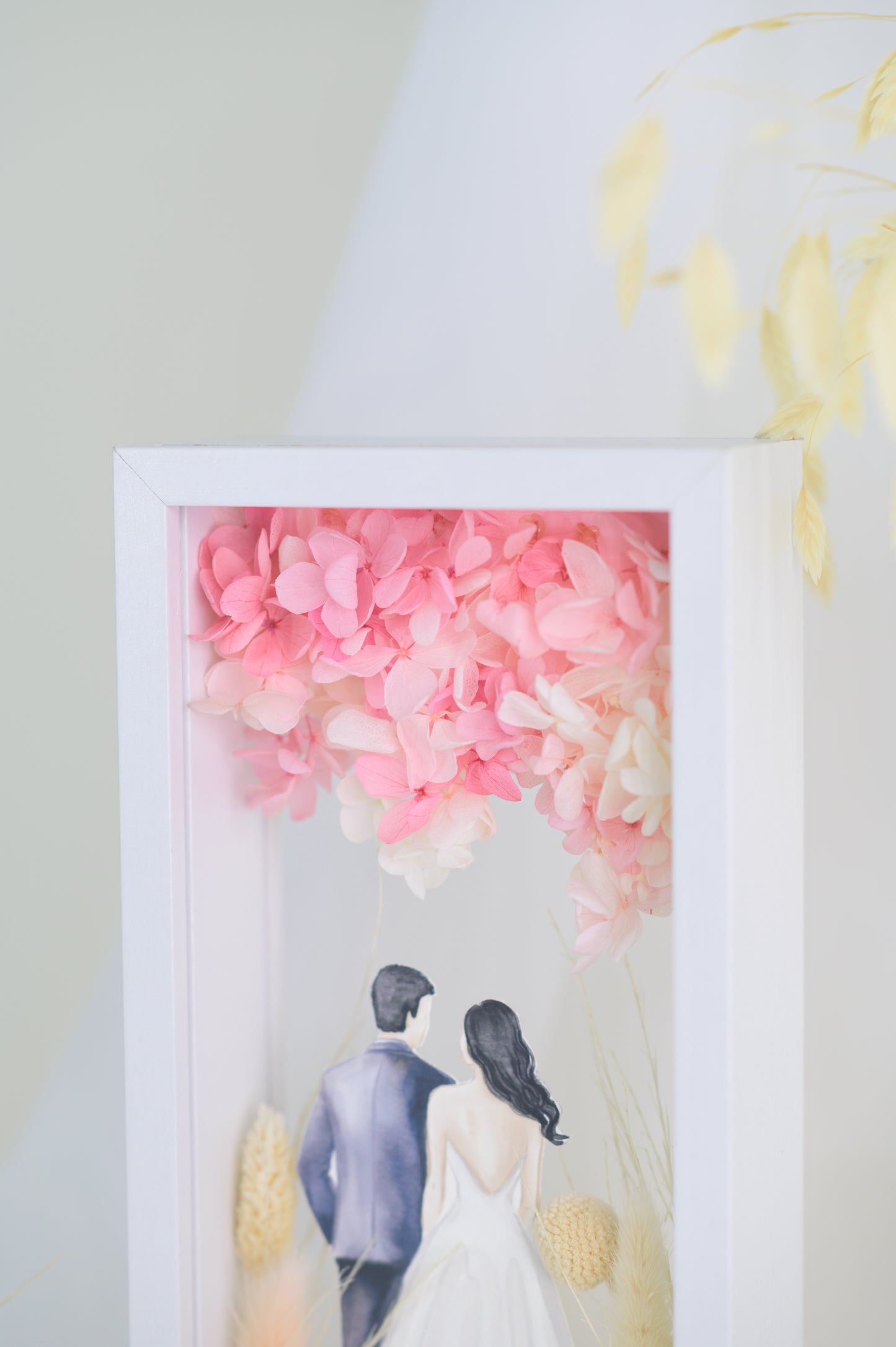 A custom wedding keepsake frame featuring a couple illustration in wedding attire, surrounded by preserved flowers in soft pinks and yellows. The frame has a front and back view, offering a beautiful display of the artwork. Customizable with names and date for a personal touch.