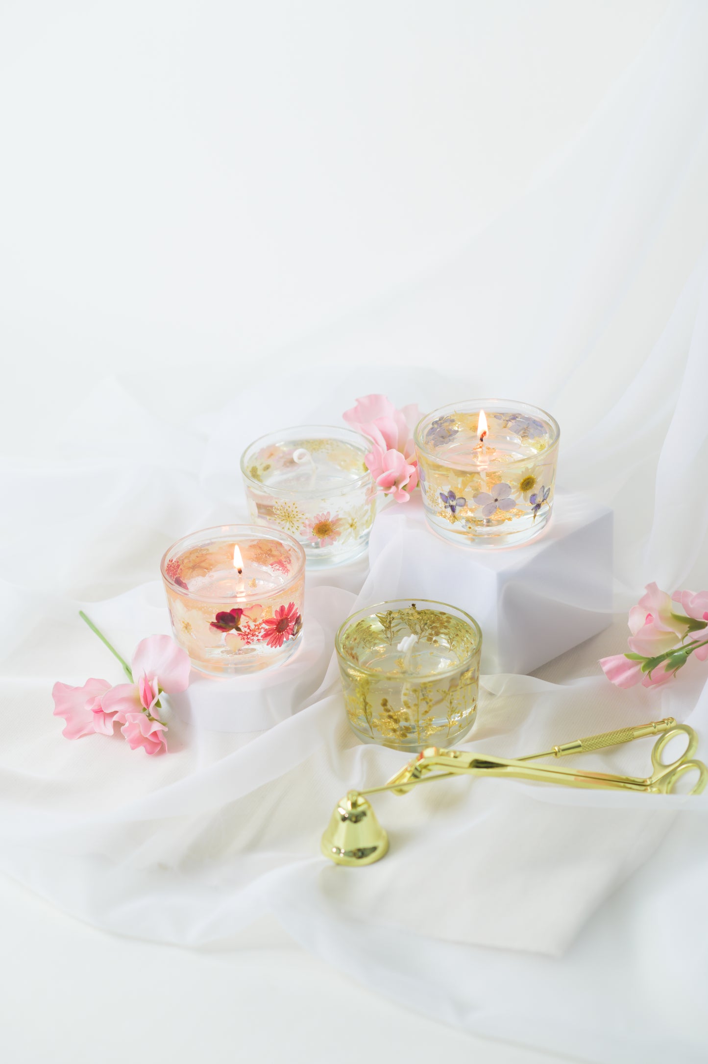 Handcrafted floral gel wax candles with embedded dried flowers and gold foil accents, burning gently on a soft fabric surface with a candle snuffer nearby.