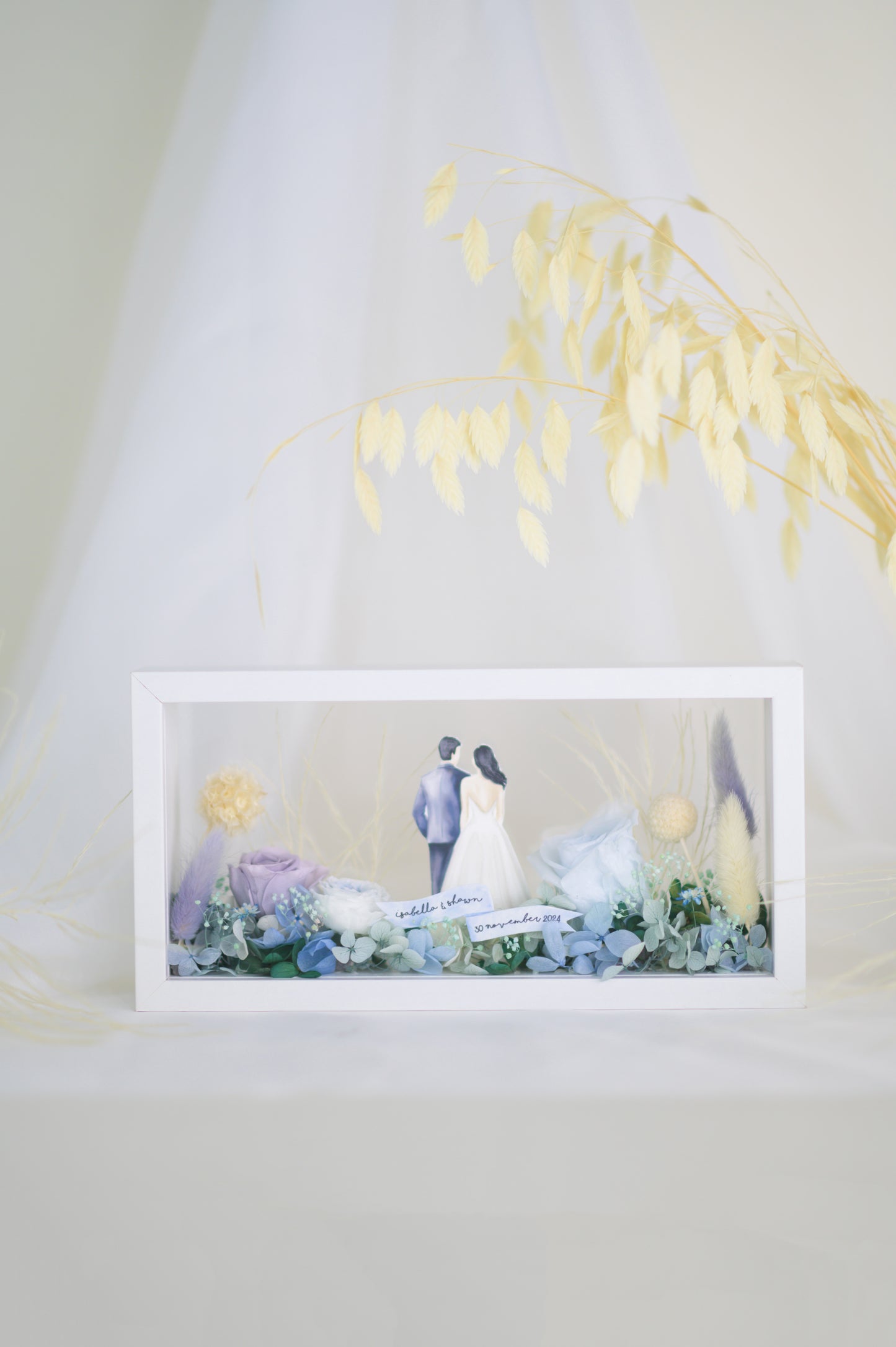 A handcrafted wedding-themed frame featuring a bride and groom with preserved flowers in soft lavender, blue, and yellow tones, displayed in a double-sided glass frame for a 360-degree view