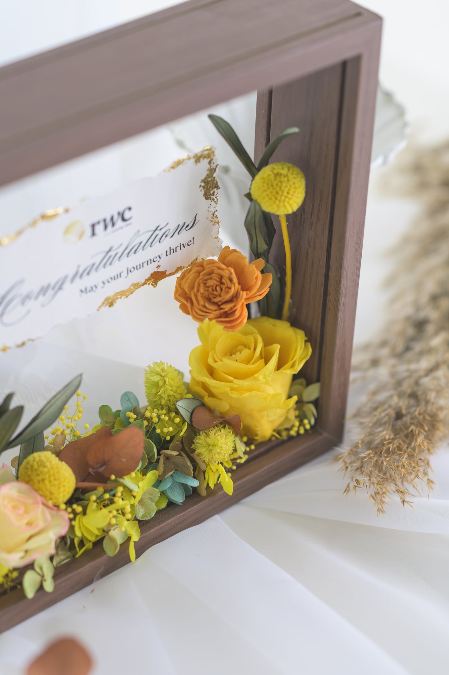 Elegant preserved flower frame with a congratulatory message, adorned with a yellow ribbon and vibrant floral arrangement, designed for a special occasion.