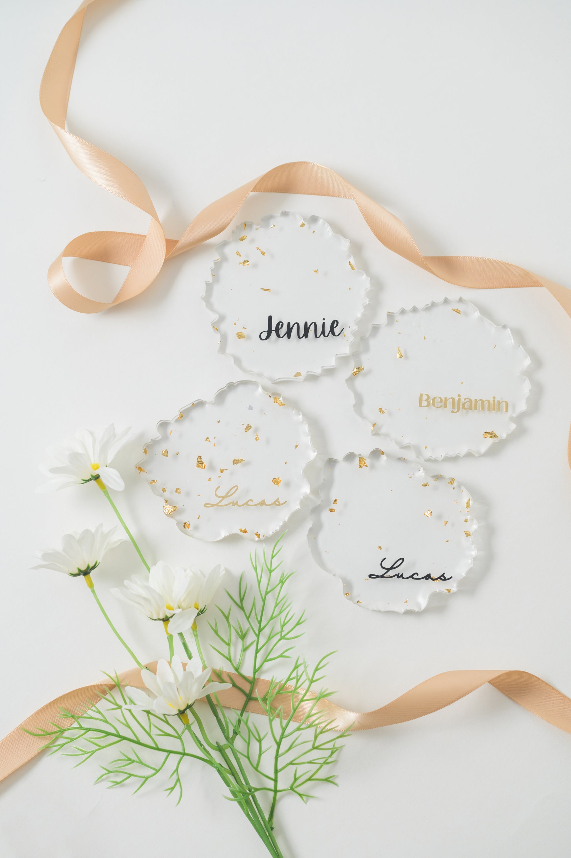 A clear, irregular-shaped resin Coaster with gold foil accents, featuring the name 'Lucas' , ' Jennie' , 'Benjamin' in elegant calligraphy, displayed alongside white daisies for a fresh, sophisticated look.
