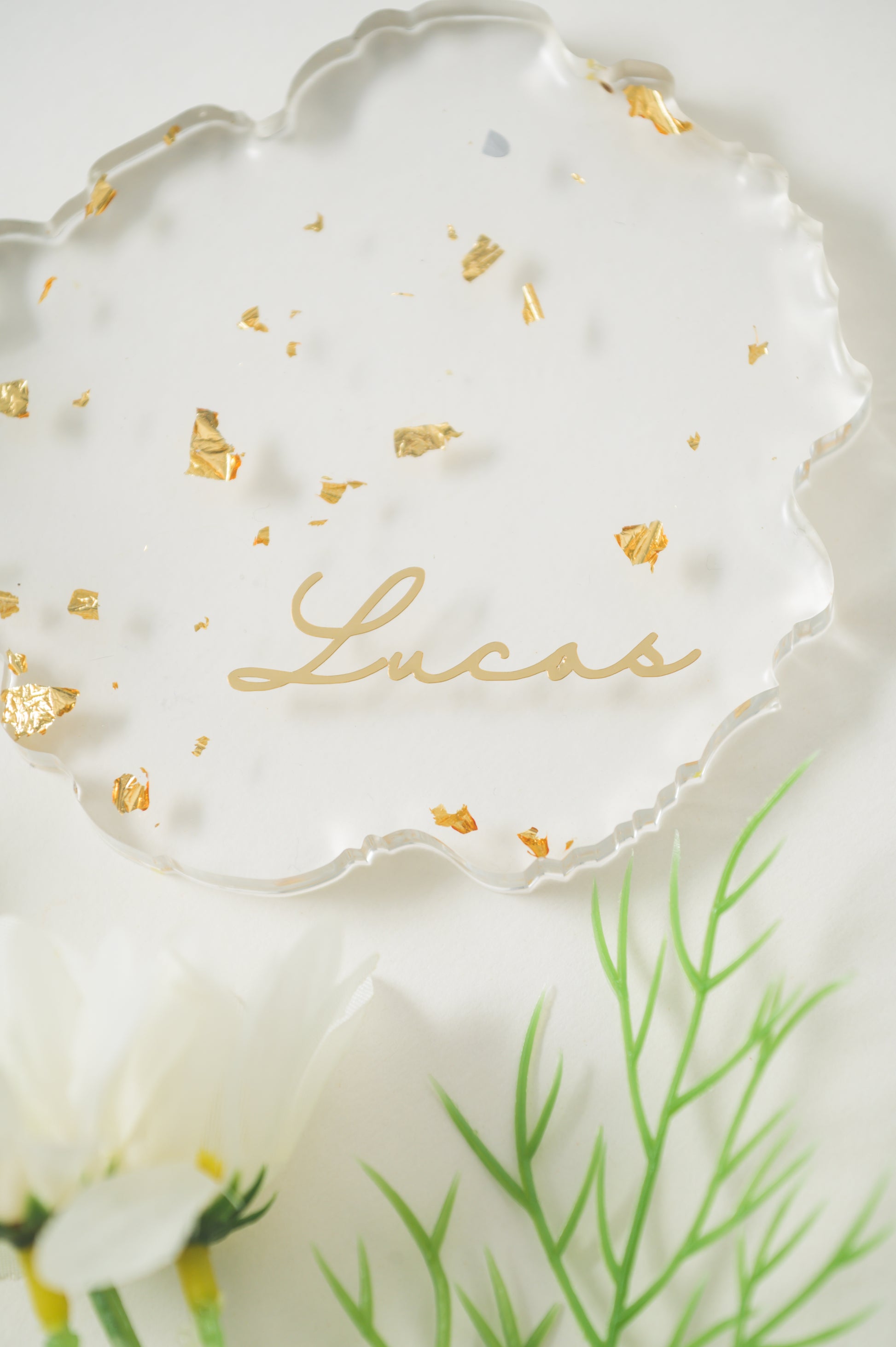 A clear, irregular-shaped resin Coaster with gold foil accents, featuring the name 'Lucas' in elegant calligraphy, displayed alongside white daisies for a fresh, sophisticated look.