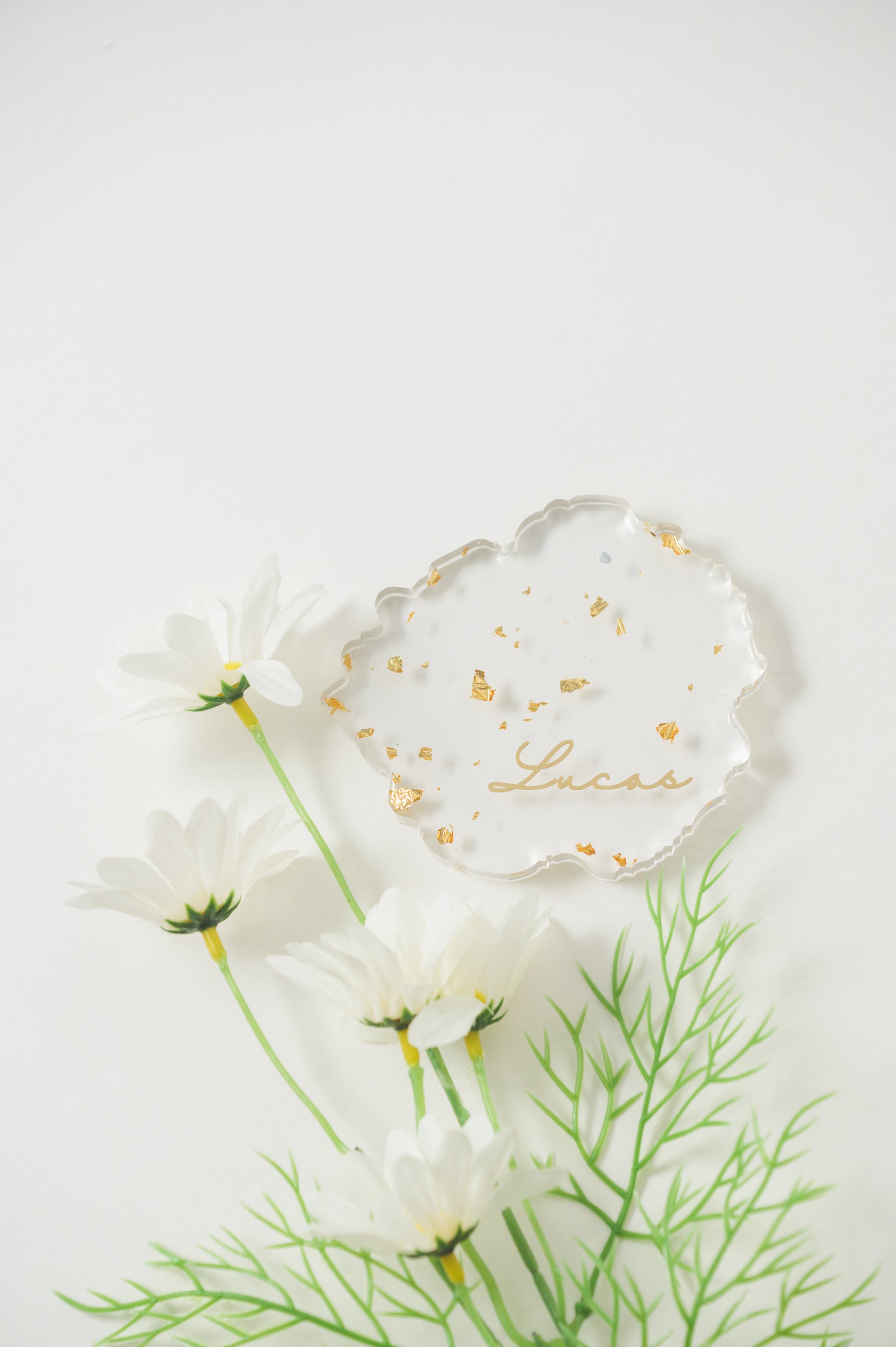 A clear, irregular-shaped resin Coaster with gold foil accents, featuring the name 'Lucas' in elegant calligraphy, displayed alongside white daisies for a fresh, sophisticated look.