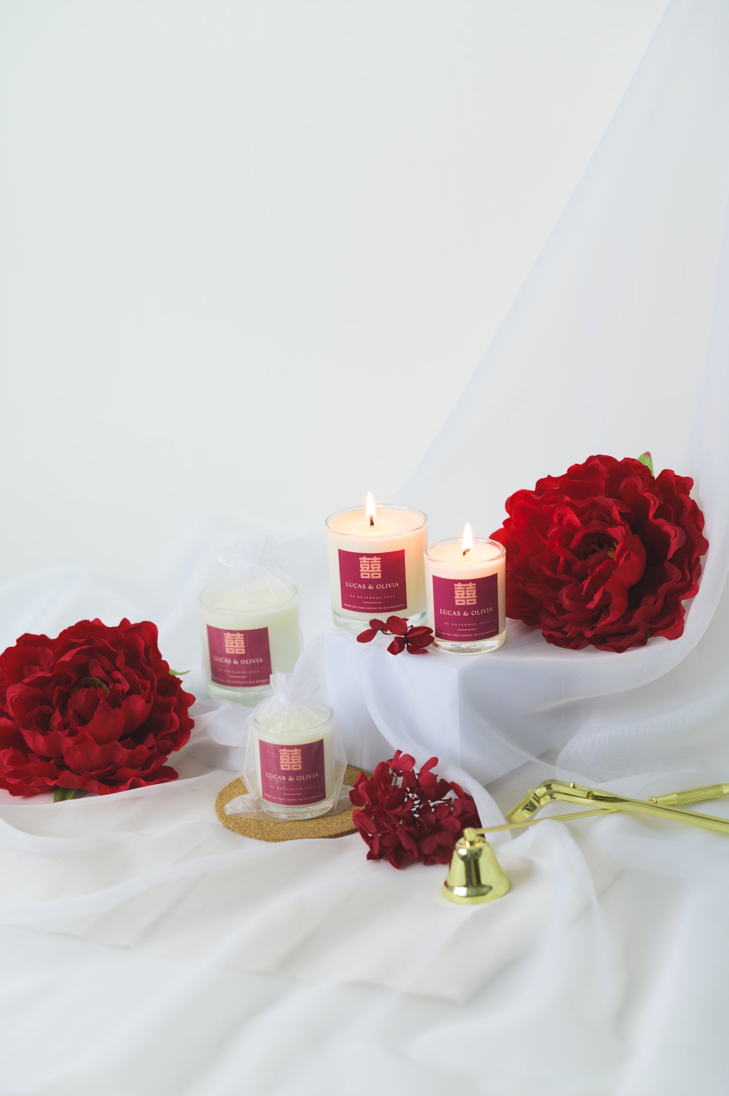 A lit custom wedding scented candle featuring a red 'Double Happiness' label with the names 'Lucas & Olivia,' wedding date, and a thank you message, placed beside red flowers as a wedding favor or gift.