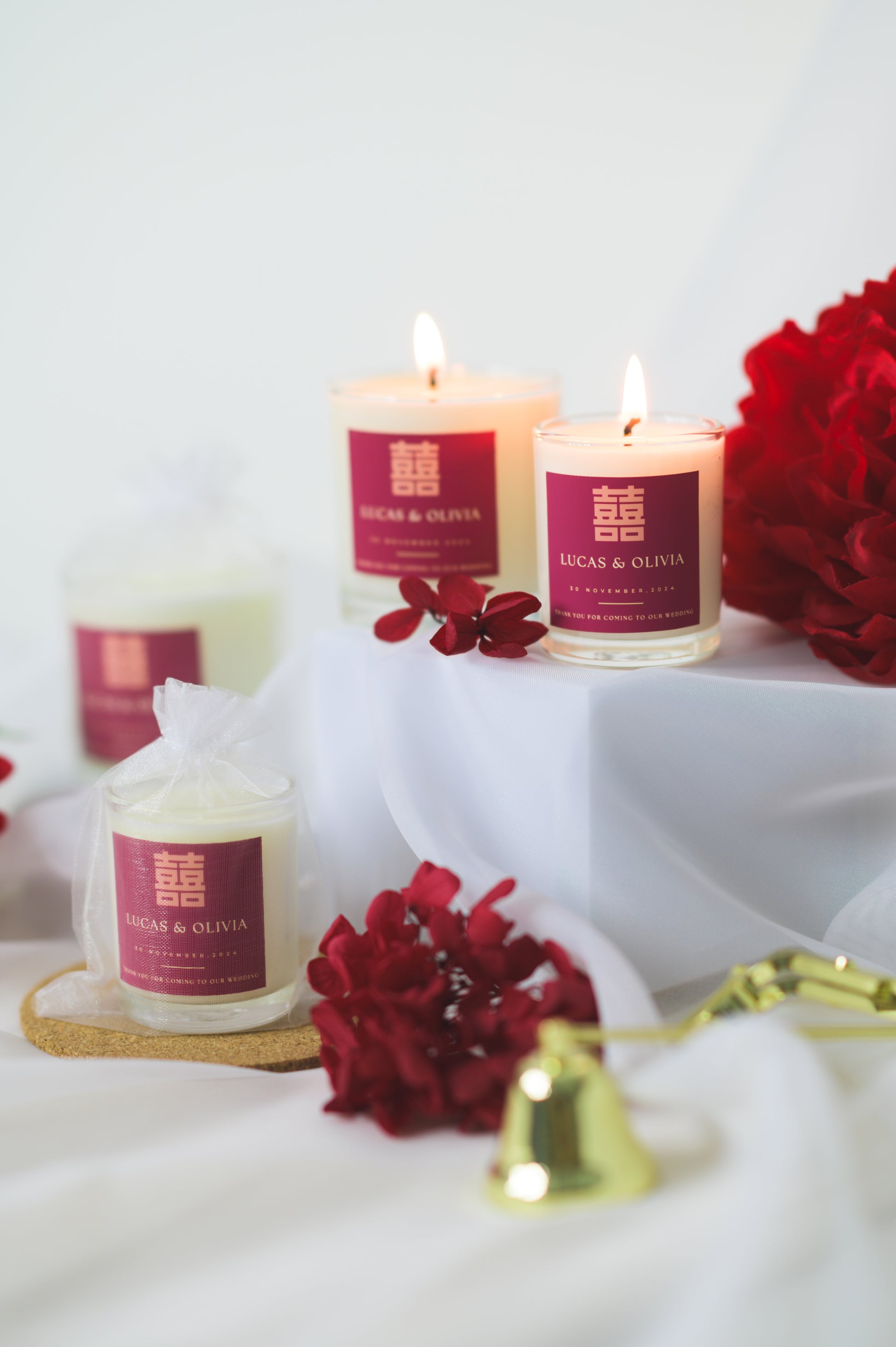 A lit custom wedding scented candle featuring a red 'Double Happiness' label with the names 'Lucas & Olivia,' wedding date, and a thank you message, placed beside red flowers as a wedding favor or gift.