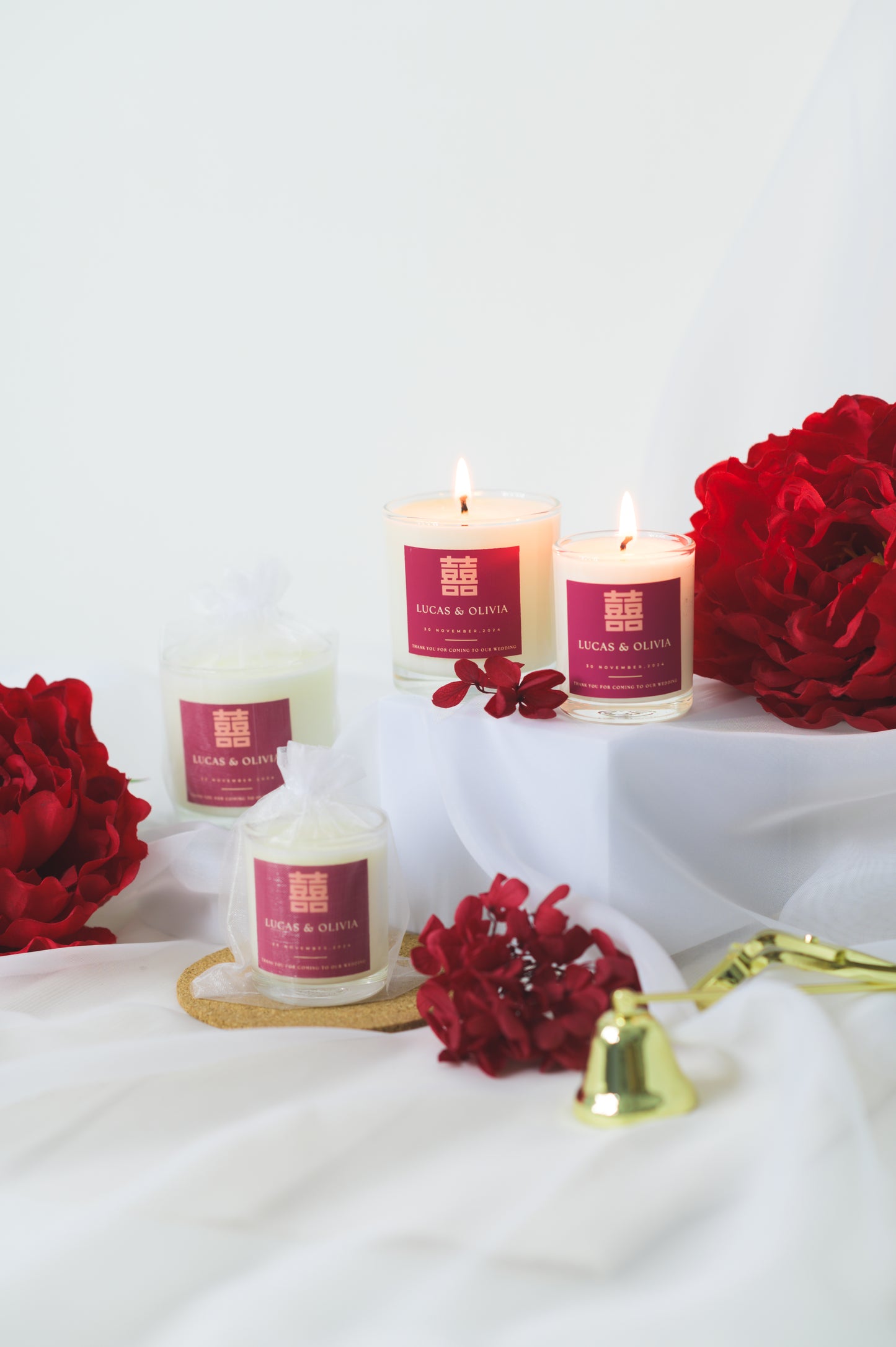 A lit custom wedding scented candle featuring a red 'Double Happiness' label with the names 'Lucas & Olivia,' wedding date, and a thank you message, placed beside red flowers as a wedding favor or gift.