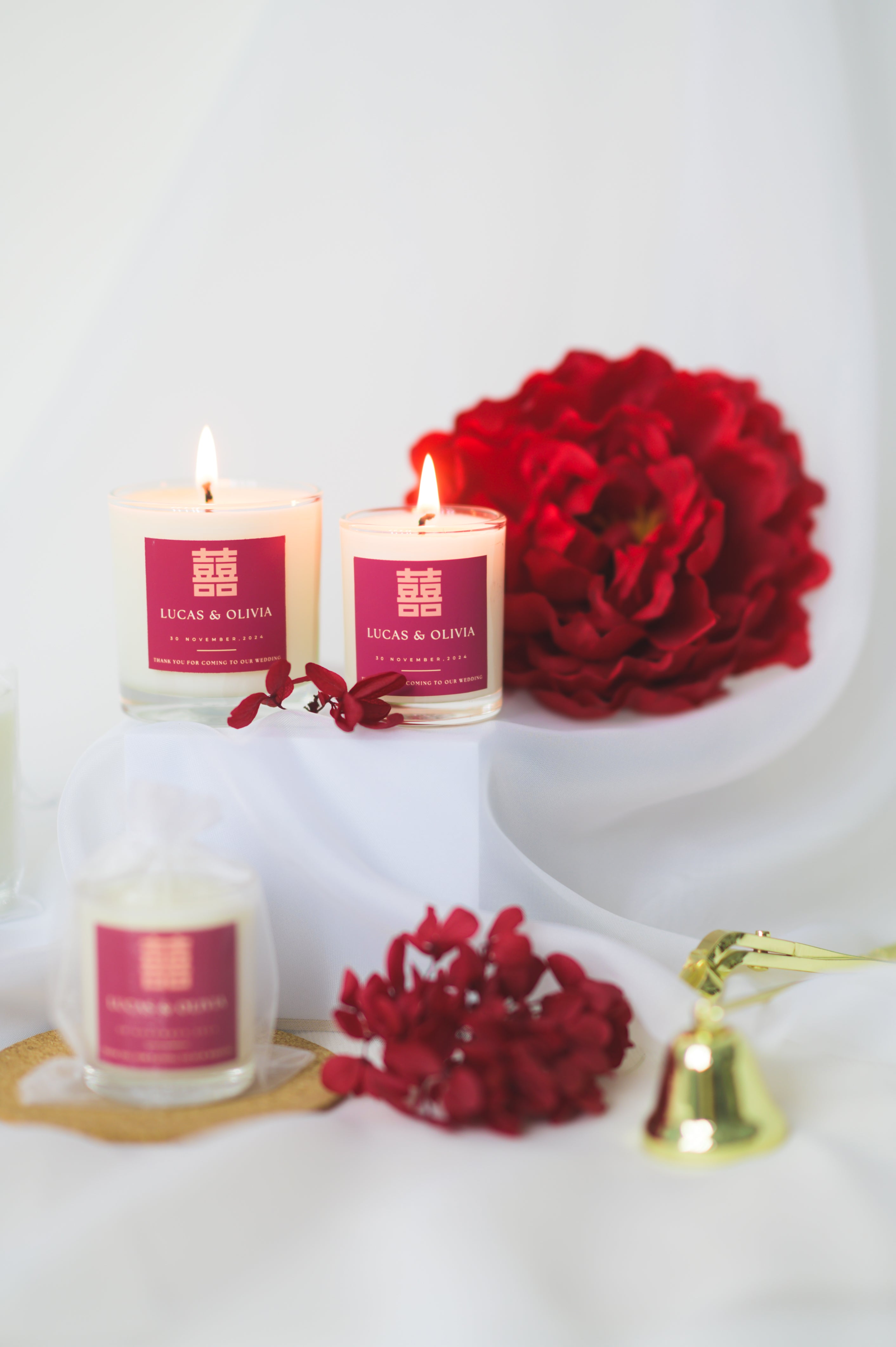 A lit custom wedding scented candle featuring a red 'Double Happiness' label with the names 'Lucas & Olivia,' wedding date, and a thank you message, placed beside red flowers as a wedding favor or gift.