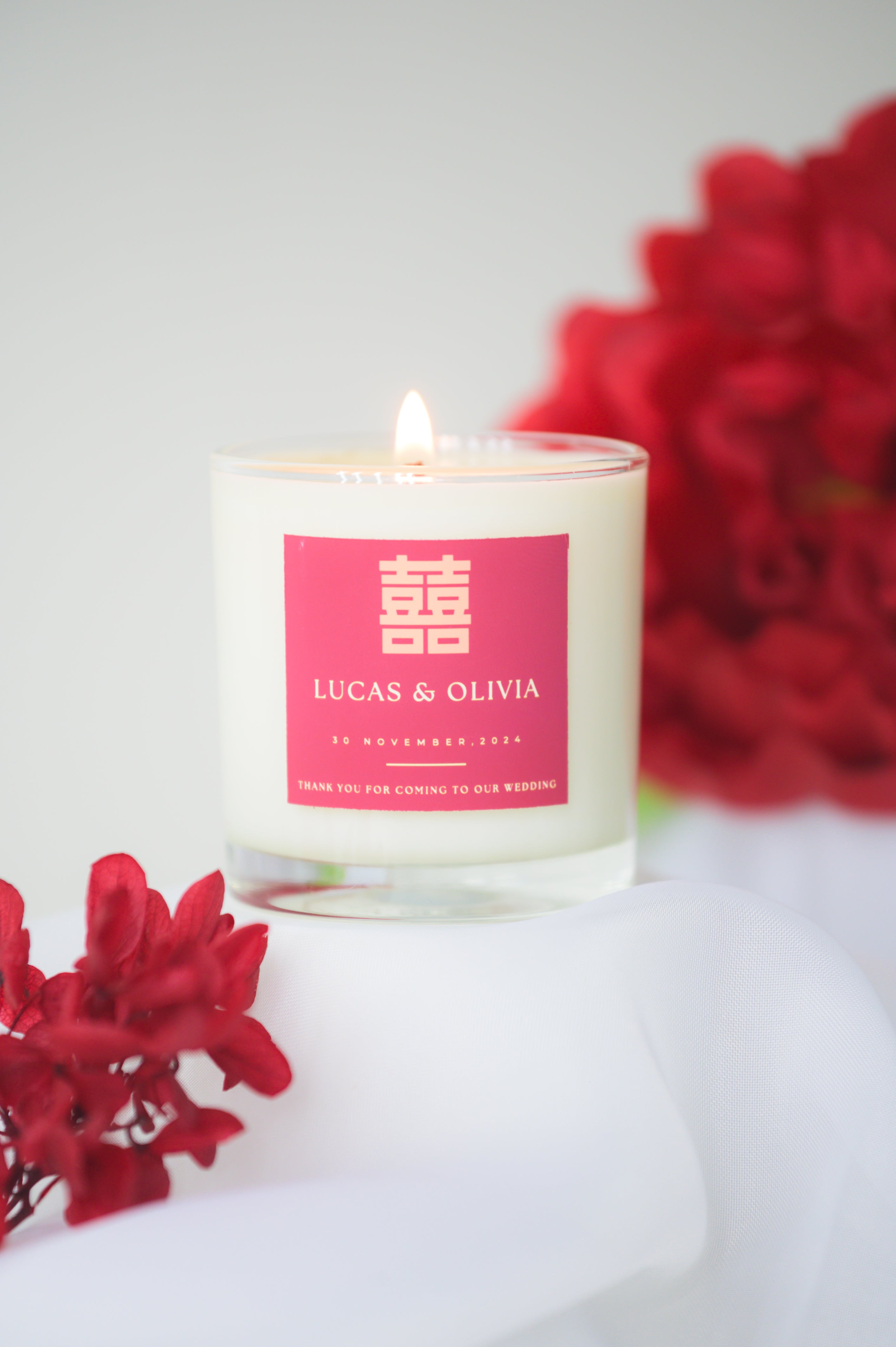 A lit custom wedding scented candle featuring a red 'Double Happiness' label with the names 'Lucas & Olivia,' wedding date, and a thank you message, placed beside red flowers as a wedding favor or gift.