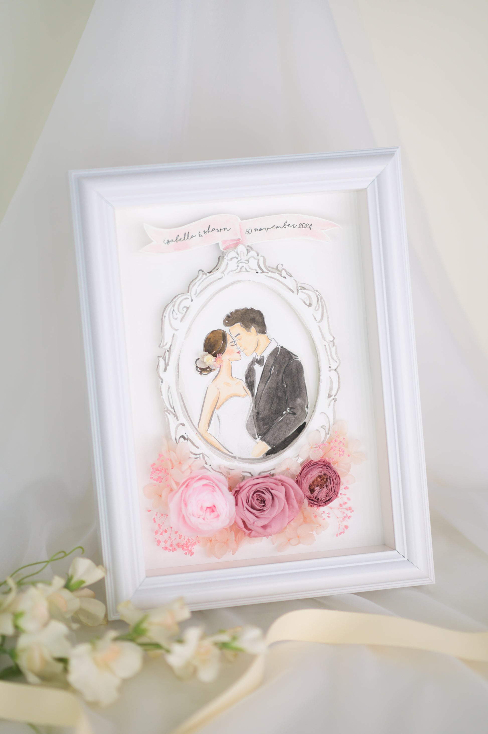 A beautifully crafted wedding frame featuring a romantic illustration of a bride and groom encased in an ornate white frame, surrounded by preserved pink roses and floral accents, personalized with names and a wedding date.