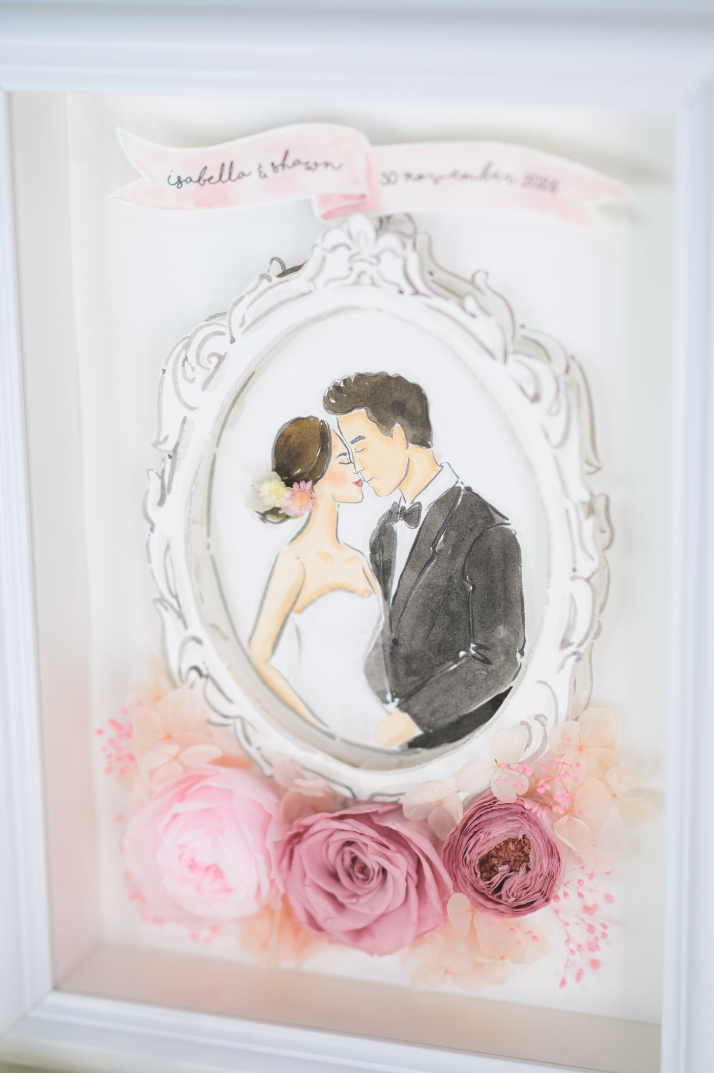A beautifully crafted wedding frame featuring a romantic illustration of a bride and groom encased in an ornate white frame, surrounded by preserved pink roses and floral accents, personalized with names and a wedding date.