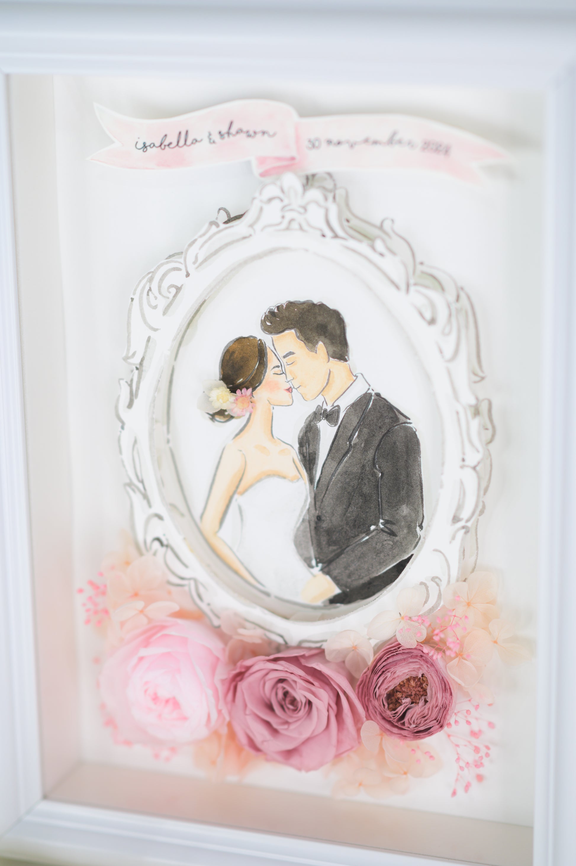 A beautifully crafted wedding frame featuring a romantic illustration of a bride and groom encased in an ornate white frame, surrounded by preserved pink roses and floral accents, personalized with names and a wedding date.