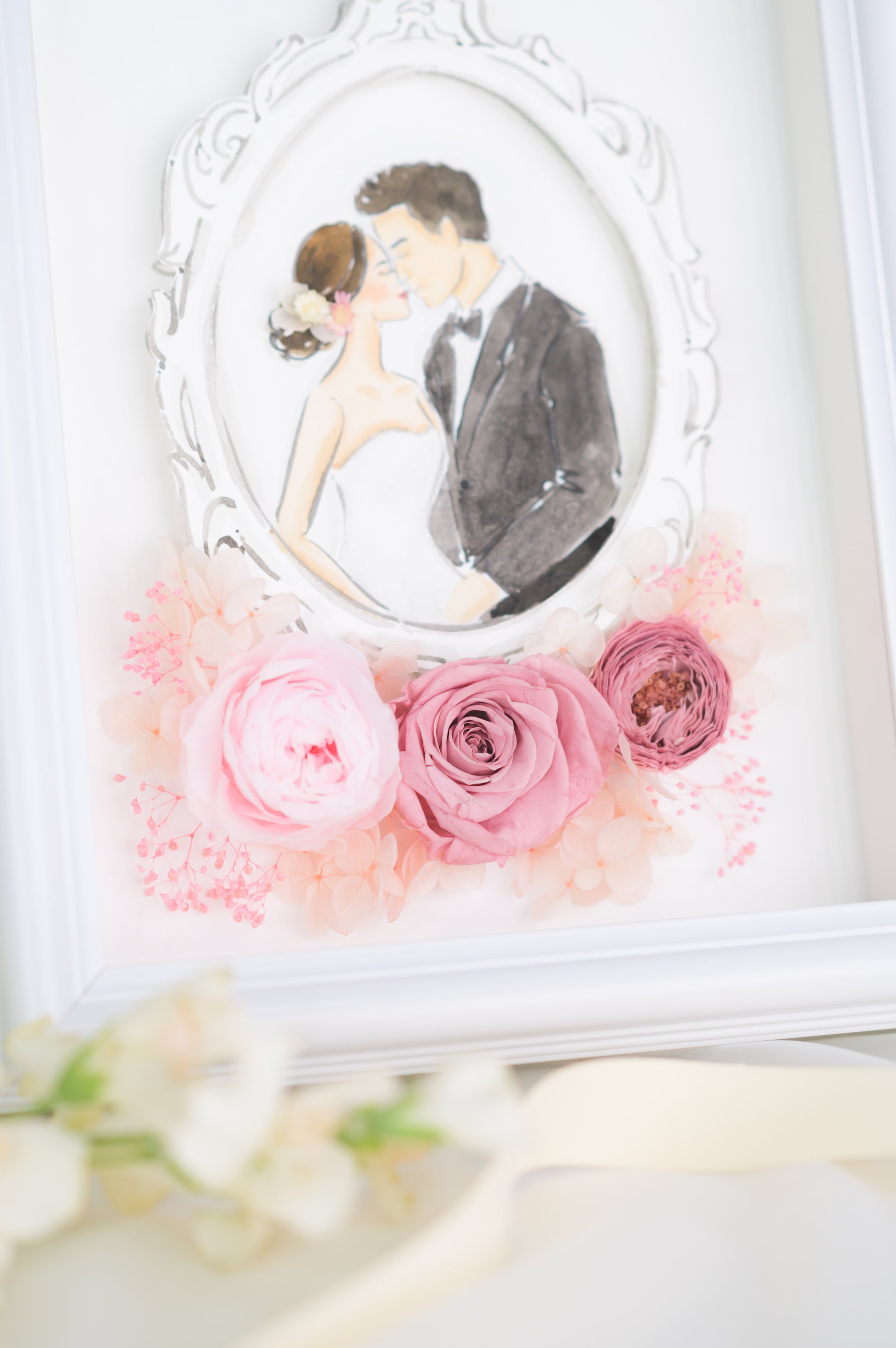 A beautifully crafted wedding frame featuring a romantic illustration of a bride and groom encased in an ornate white frame, surrounded by preserved pink roses and floral accents, personalized with names and a wedding date.