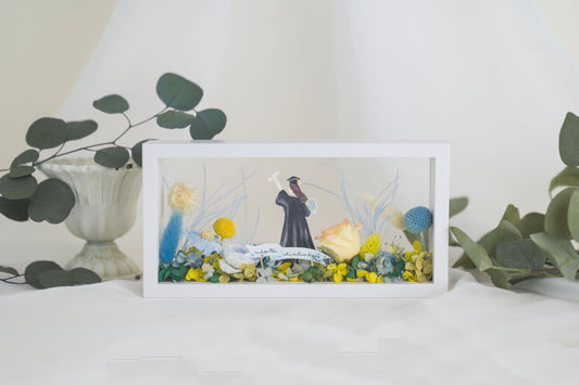 Graduation-themed preserved flower frame featuring a graduate figure holding a diploma, surrounded by vibrant yellow, blue, and white blooms.