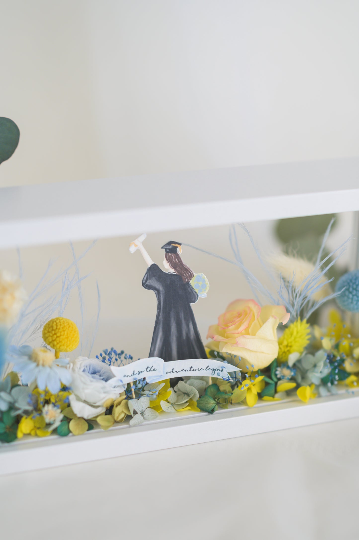 Graduation-themed preserved flower frame featuring a graduate figure holding a diploma, surrounded by vibrant yellow, blue, and white blooms.