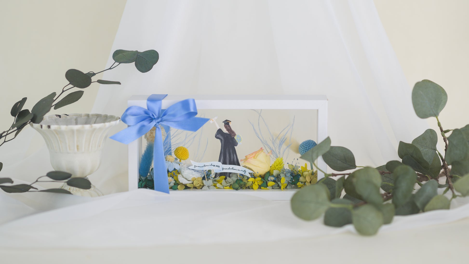 Graduation-themed preserved flower frame featuring a graduate figure holding a diploma, surrounded by vibrant yellow, blue, and white blooms.