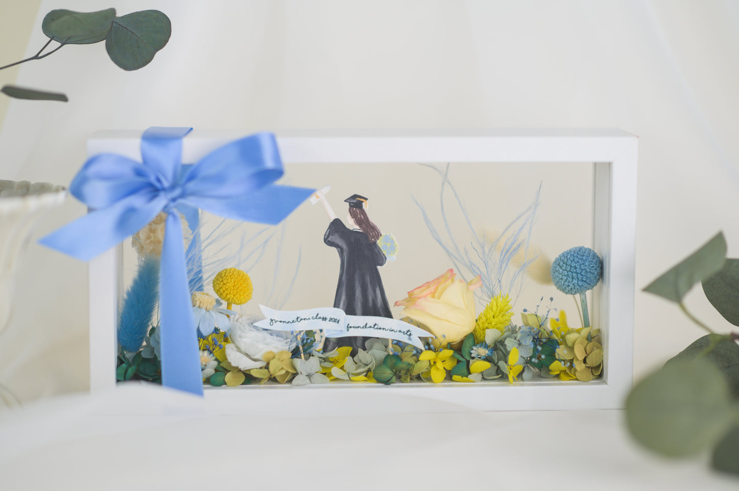 Graduation-themed preserved flower frame featuring a graduate figure holding a diploma, surrounded by vibrant yellow, blue, and white blooms.