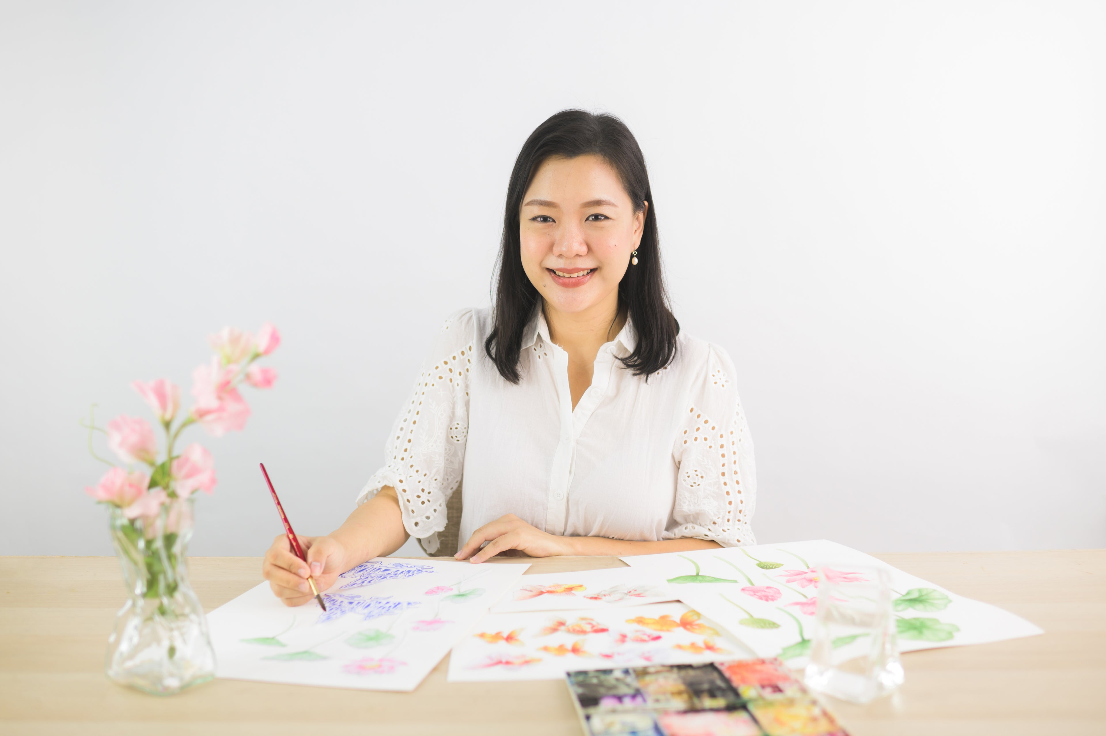 Artist Limzy working on a floral painting, seated at a table with watercolor supplies and pink flowers in a vase, wearing a white blouse and smiling as she creates her artwork.