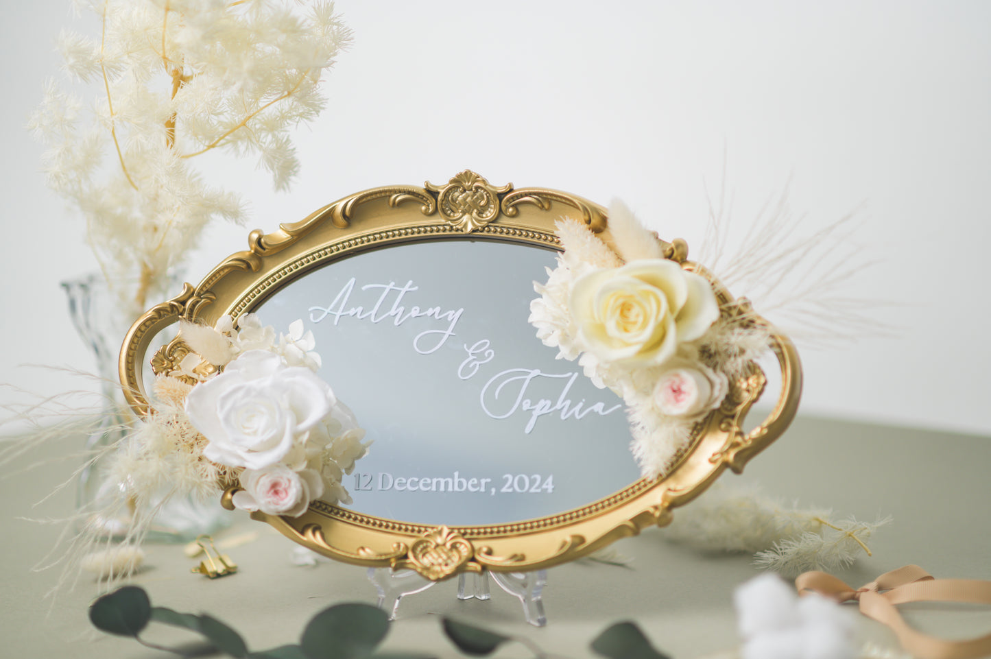 Ornate gold-framed wedding mirror featuring preserved white flowers, with customizable names Anthony & Sophia and wedding date, beautifully displayed on a reception table with floral accents.