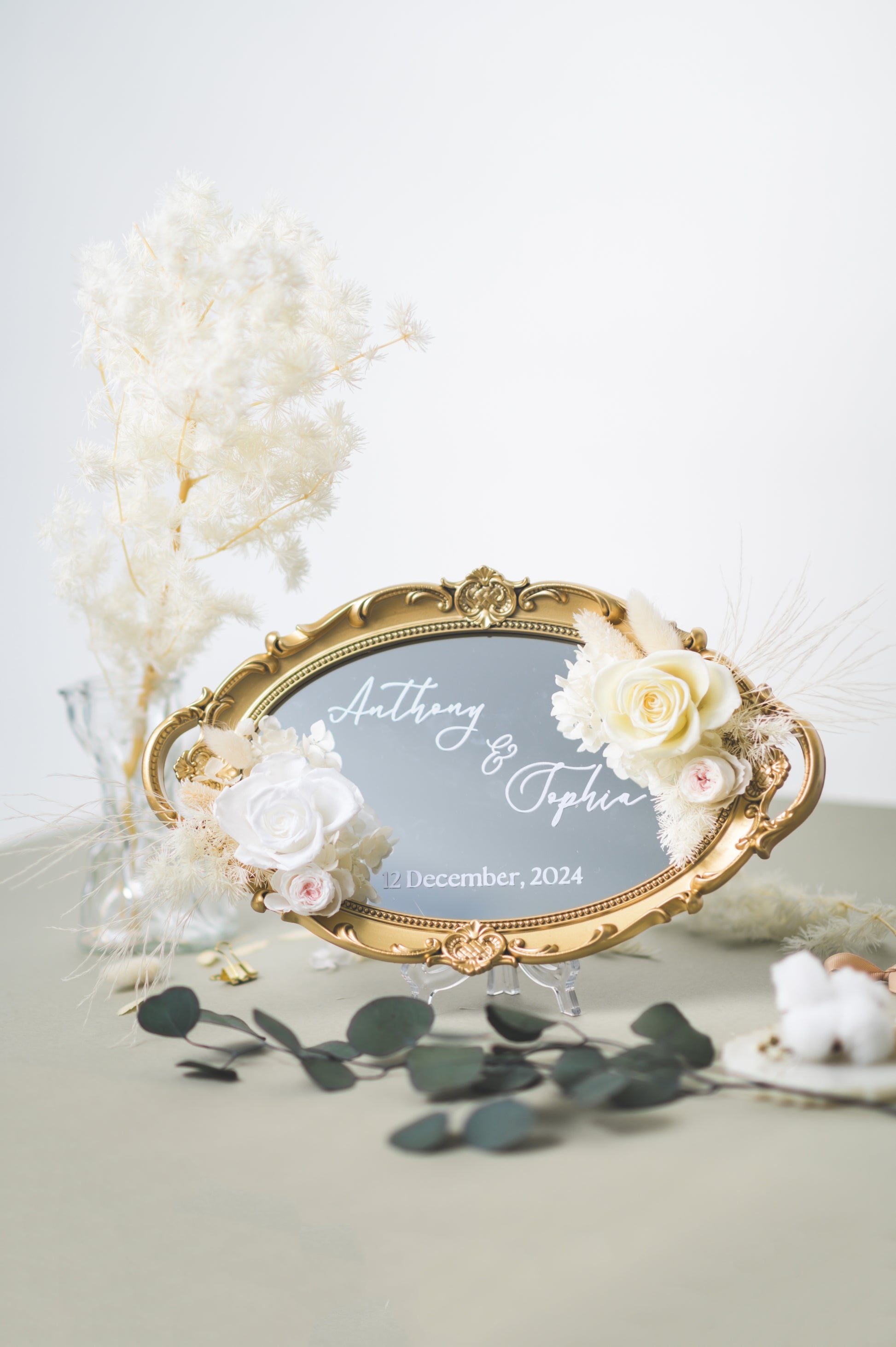 Ornate gold-framed wedding mirror featuring preserved white flowers, with customizable names Anthony & Sophia and wedding date, beautifully displayed on a reception table with floral accents.