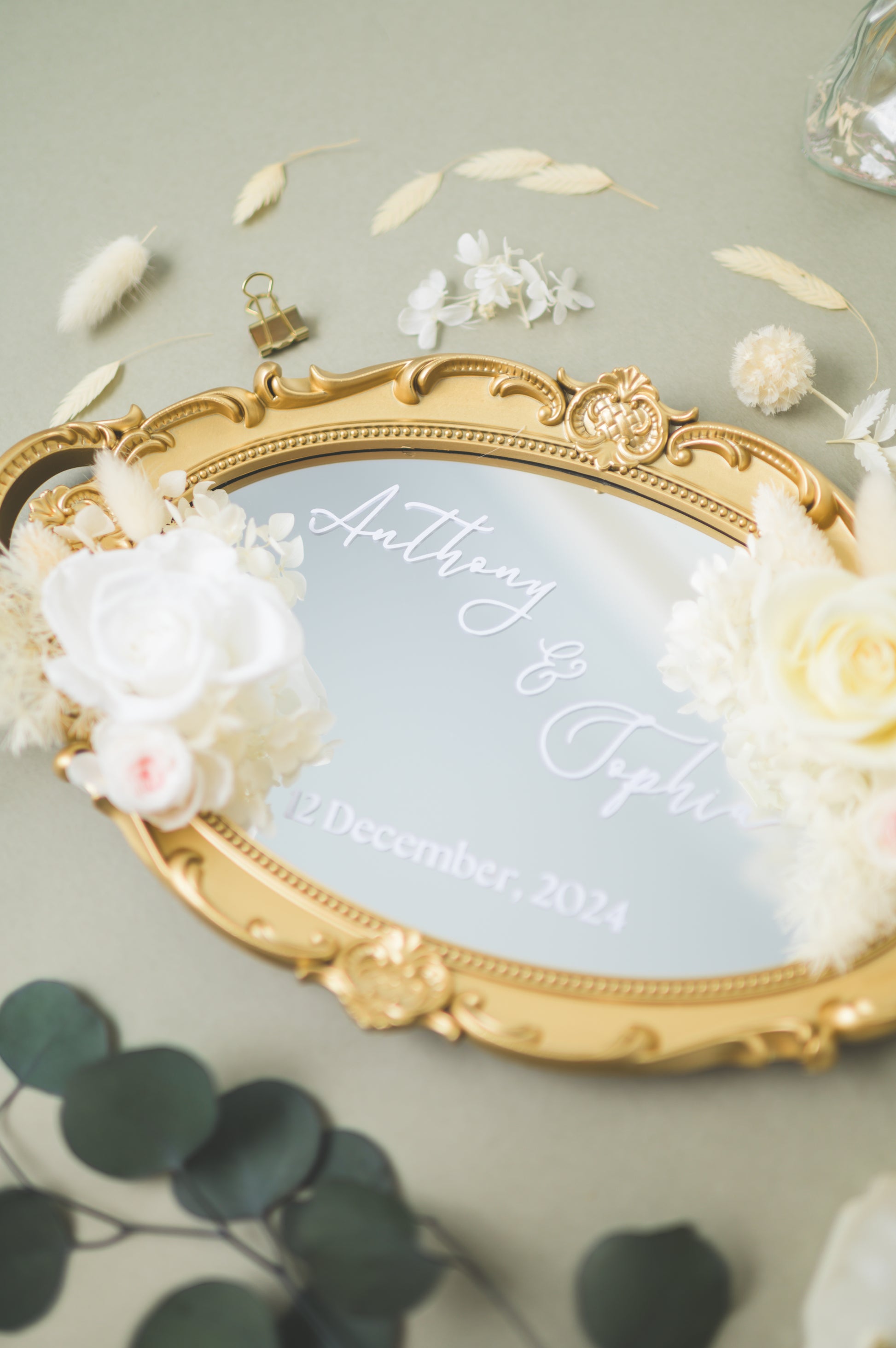 Ornate gold-framed wedding mirror featuring preserved white flowers, with customizable names Anthony & Sophia and wedding date, beautifully displayed on a reception table with floral accents.