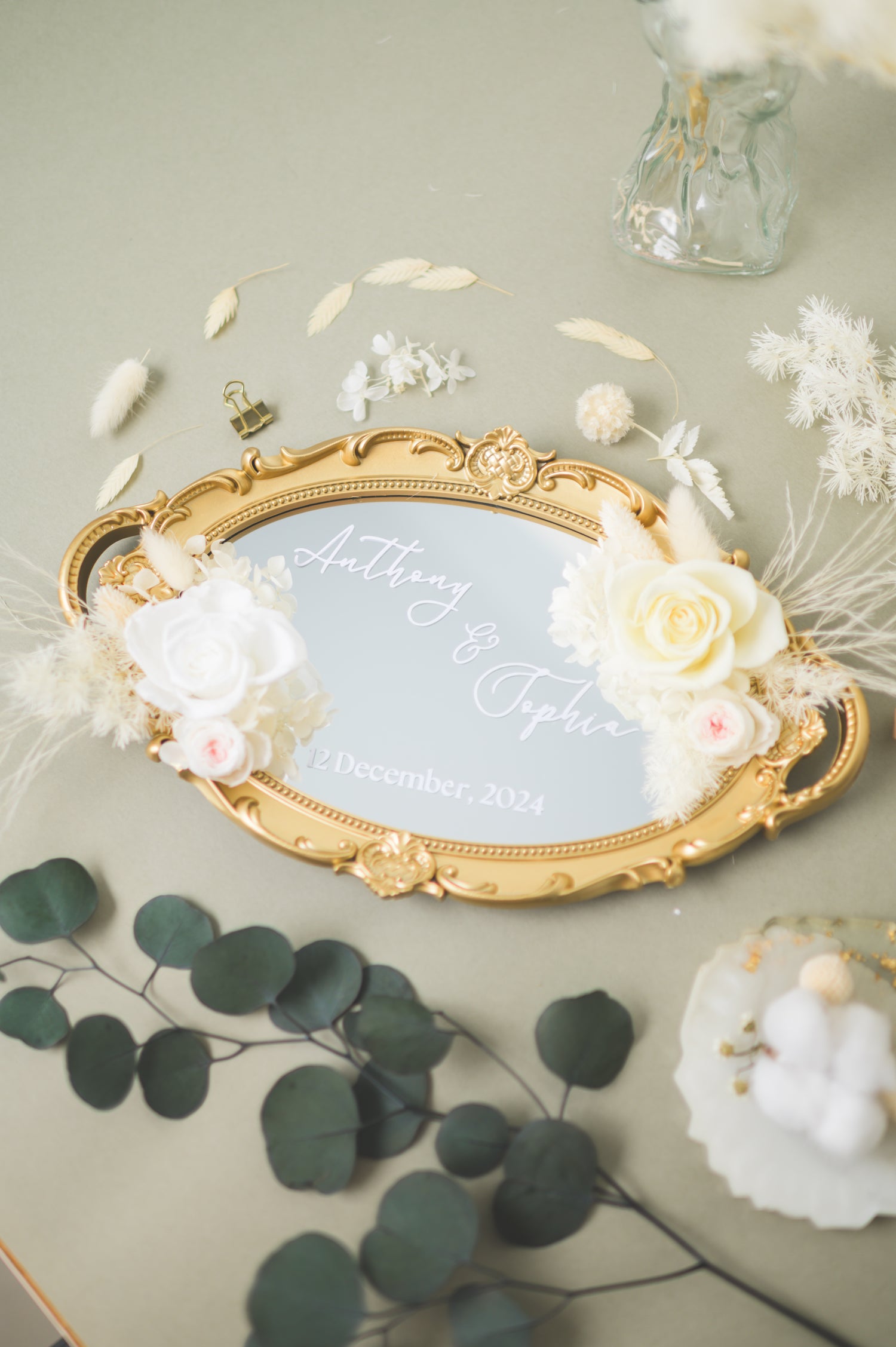 Ornate gold-framed wedding mirror featuring preserved white flowers, with customizable names Anthony & Sophia and wedding date, beautifully displayed on a reception table with floral accents.