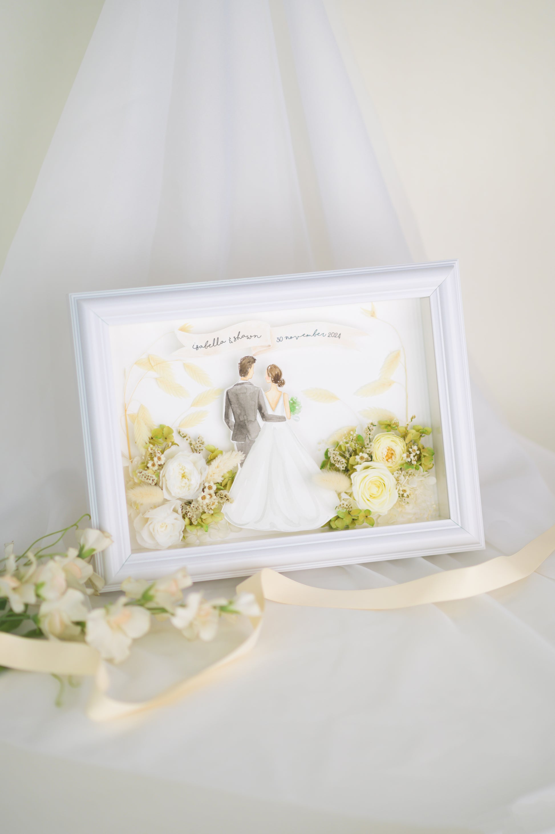 Personalized wedding frame featuring a bride and groom illustration with preserved white and green flowers, custom names and date, framed in white.