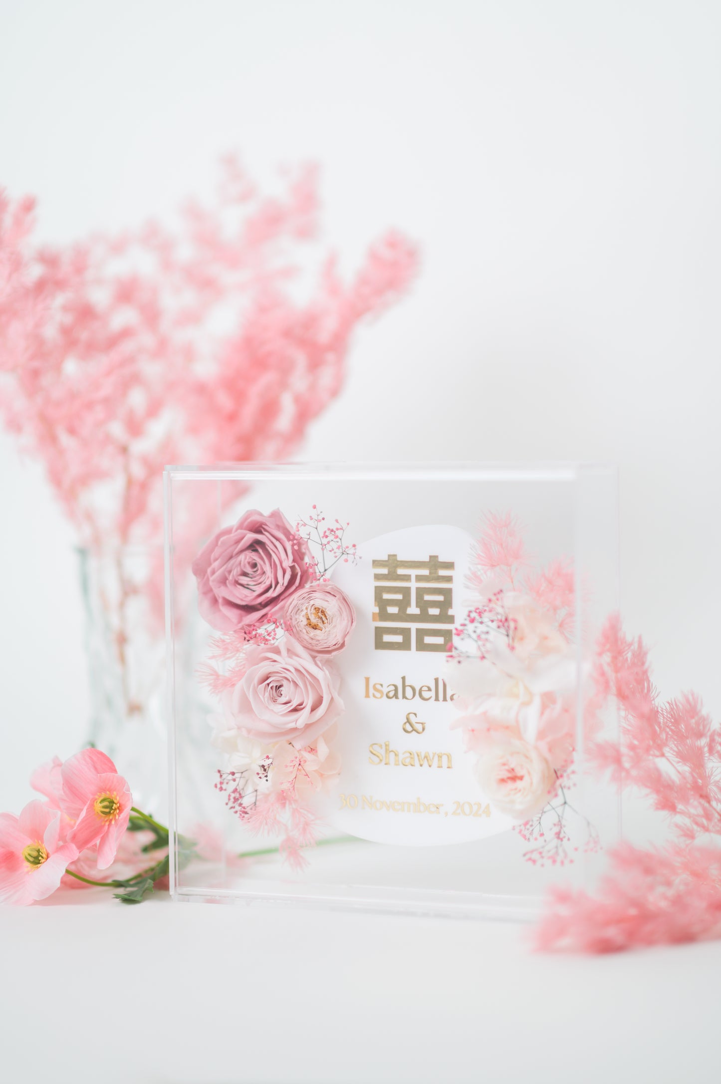 Personalized wedding frame with preserved pink flowers, featuring custom names Isabella & Shawn and a wedding date, with elegant floral design in a clear acrylic display.
