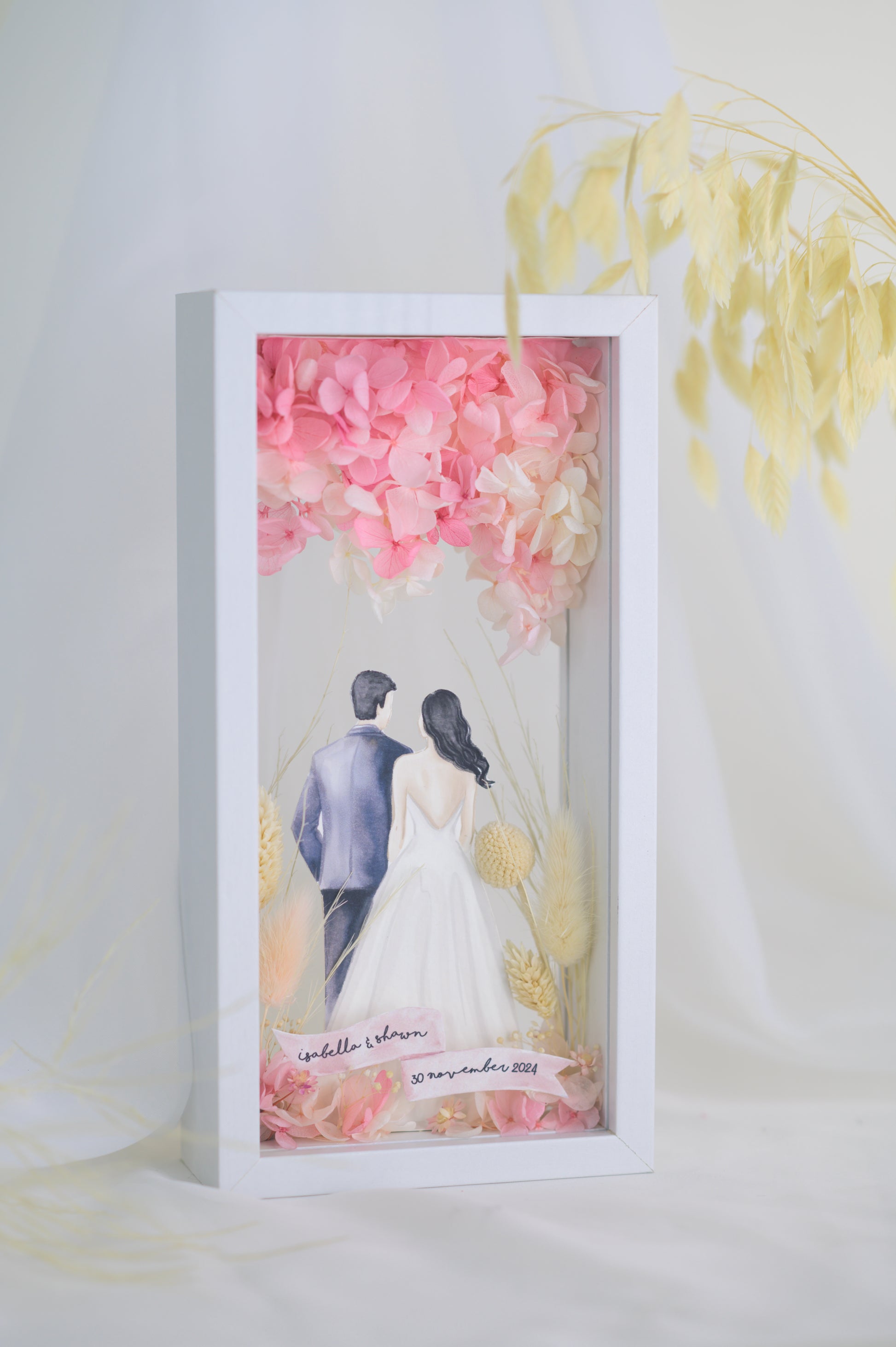 A custom wedding keepsake frame featuring a couple illustration in wedding attire, surrounded by preserved flowers in soft pinks and yellows. The frame has a front and back view, offering a beautiful display of the artwork. Customizable with names and date for a personal touch.