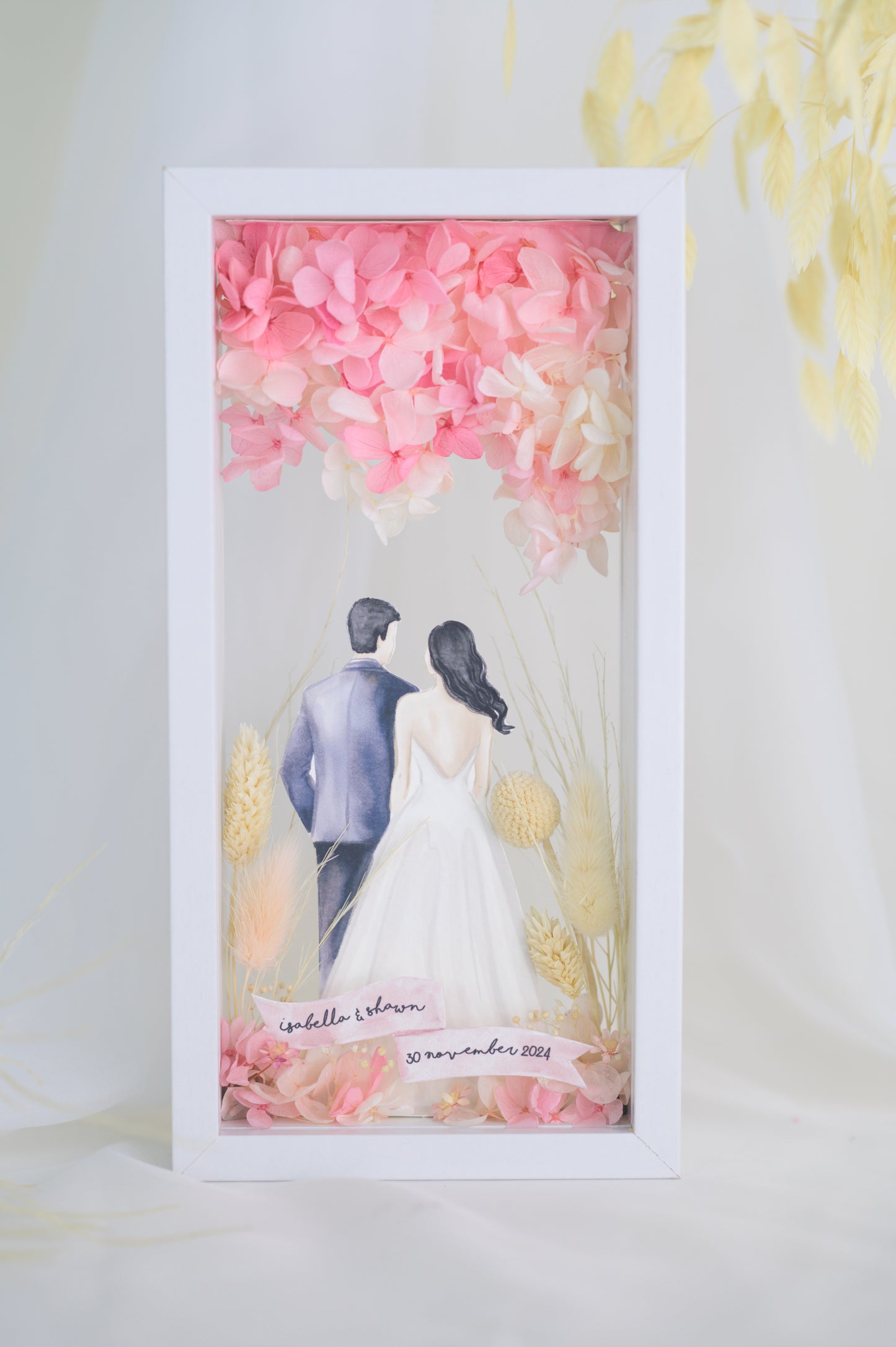 A custom wedding keepsake frame featuring a couple illustration in wedding attire, surrounded by preserved flowers in soft pinks and yellows. The frame has a front and back view, offering a beautiful display of the artwork. Customizable with names and date for a personal touch.