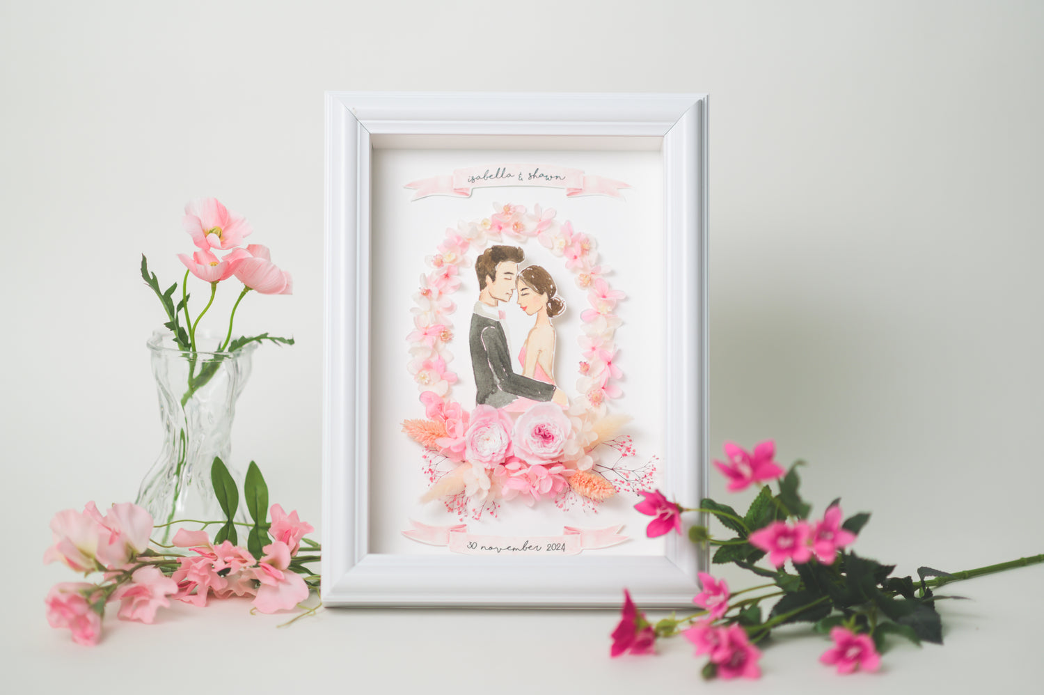 Romantic wedding frame featuring a bride and groom illustration surrounded by preserved pink flowers, with custom names and date, framed in white.