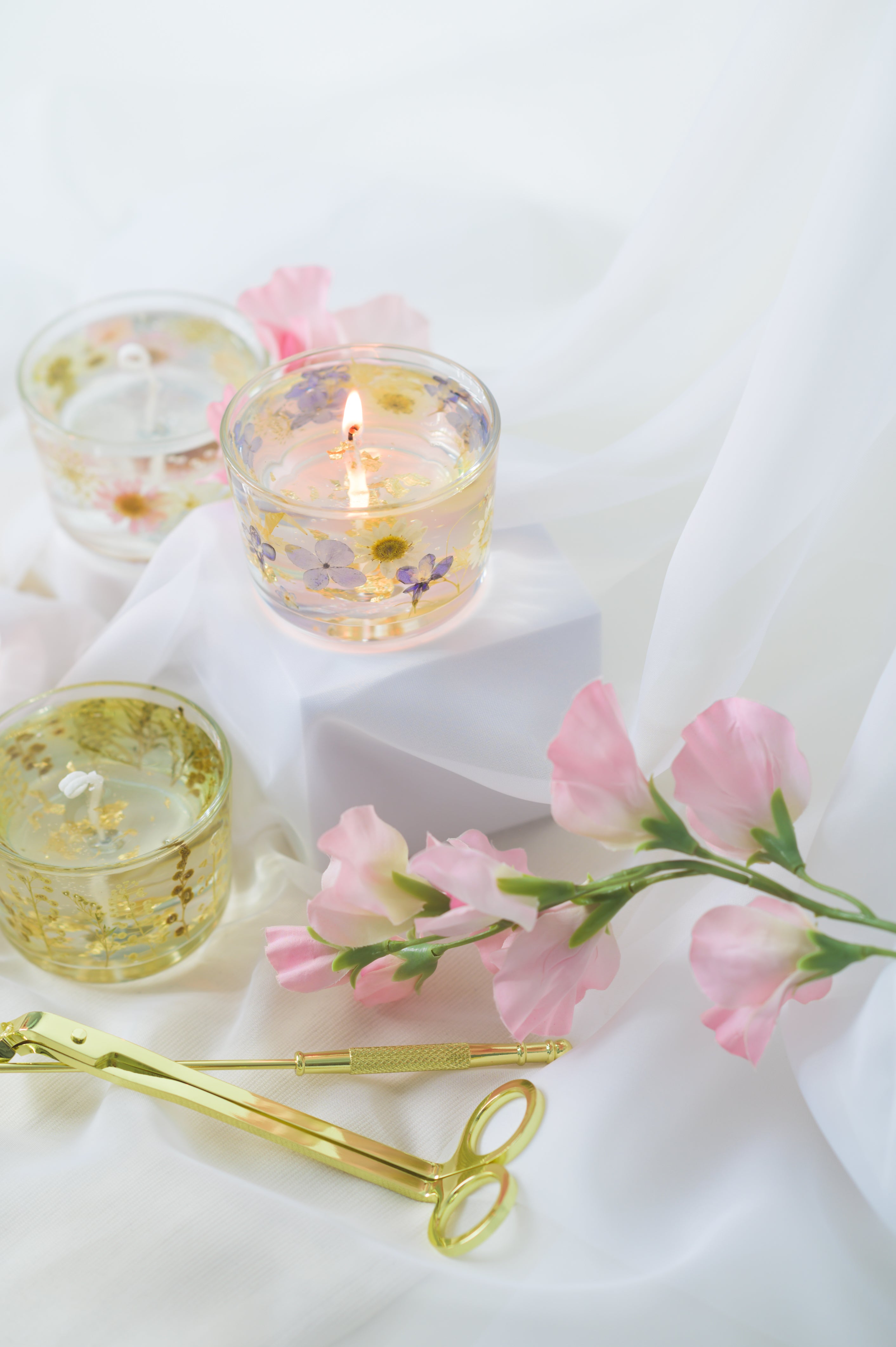 Handcrafted floral gel wax candles with embedded dried flowers and gold foil accents, burning gently on a soft fabric surface with a candle snuffer nearby.