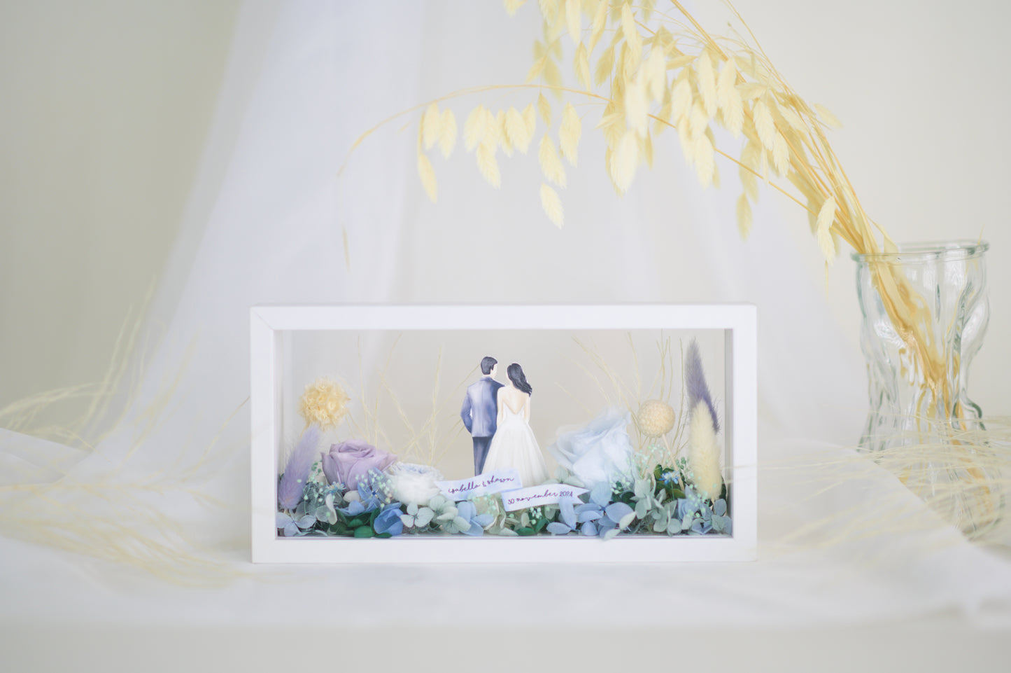 A handcrafted wedding-themed frame featuring a bride and groom with preserved flowers in soft lavender, blue, and yellow tones, displayed in a double-sided glass frame for a 360-degree view
