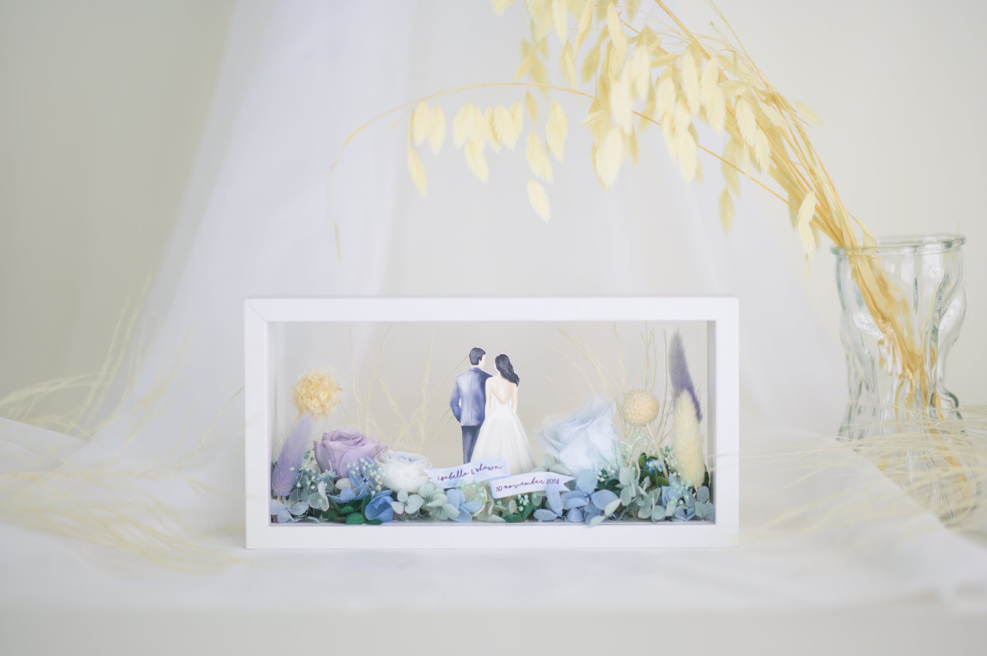 A handcrafted wedding-themed frame featuring a bride and groom with preserved flowers in soft lavender, blue, and yellow tones, displayed in a double-sided glass frame for a 360-degree view