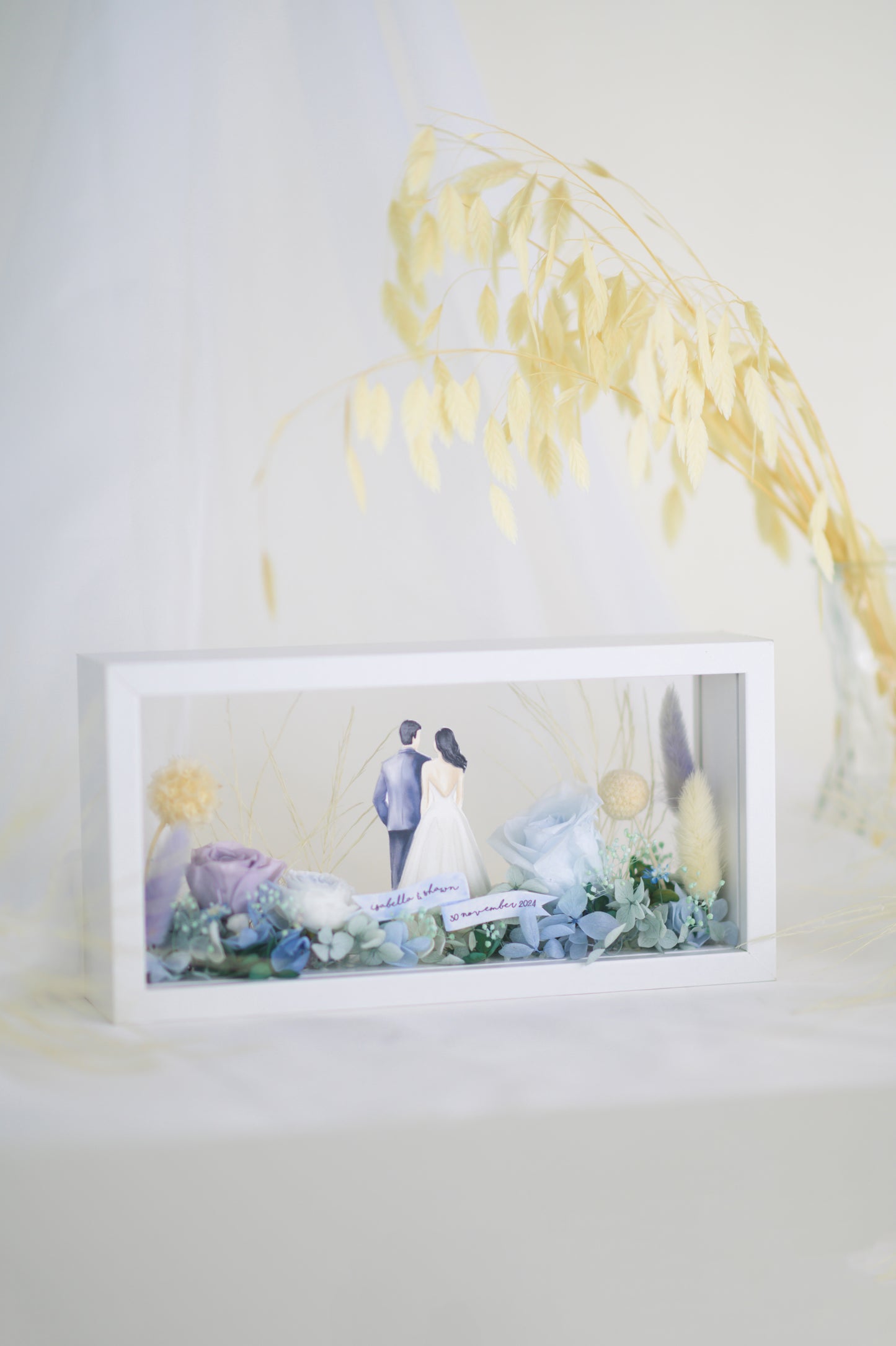 A handcrafted wedding-themed frame featuring a bride and groom with preserved flowers in soft lavender, blue, and yellow tones, displayed in a double-sided glass frame for a 360-degree view