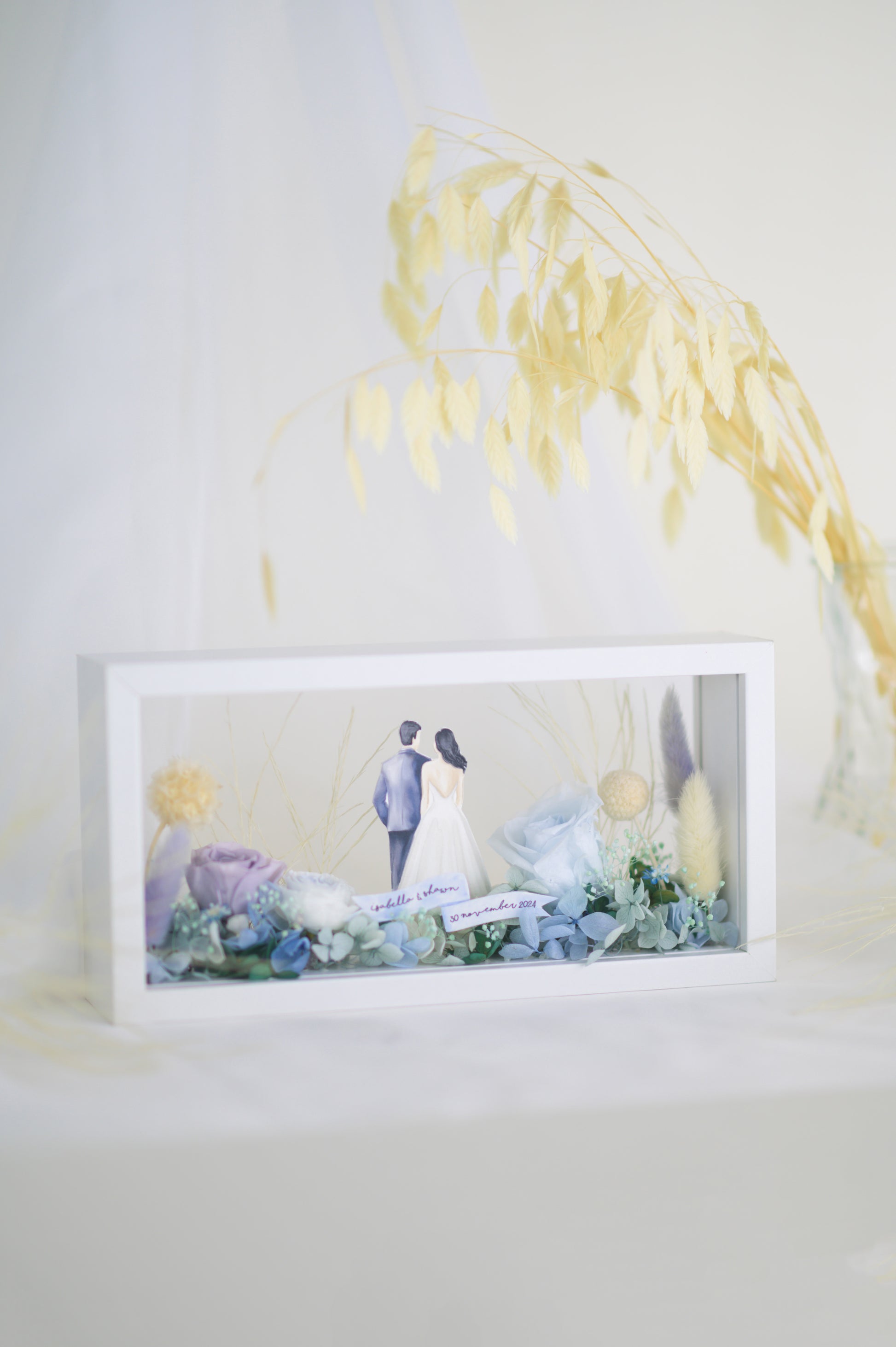 A handcrafted wedding-themed frame featuring a bride and groom with preserved flowers in soft lavender, blue, and yellow tones, displayed in a double-sided glass frame for a 360-degree view