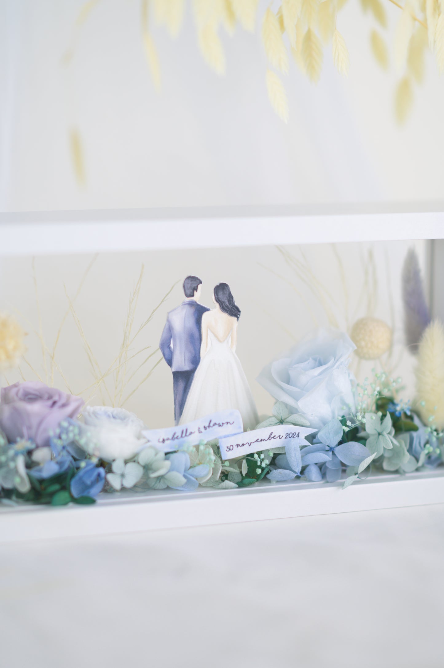 A handcrafted wedding-themed frame featuring a bride and groom with preserved flowers in soft lavender, blue, and yellow tones, displayed in a double-sided glass frame for a 360-degree view
