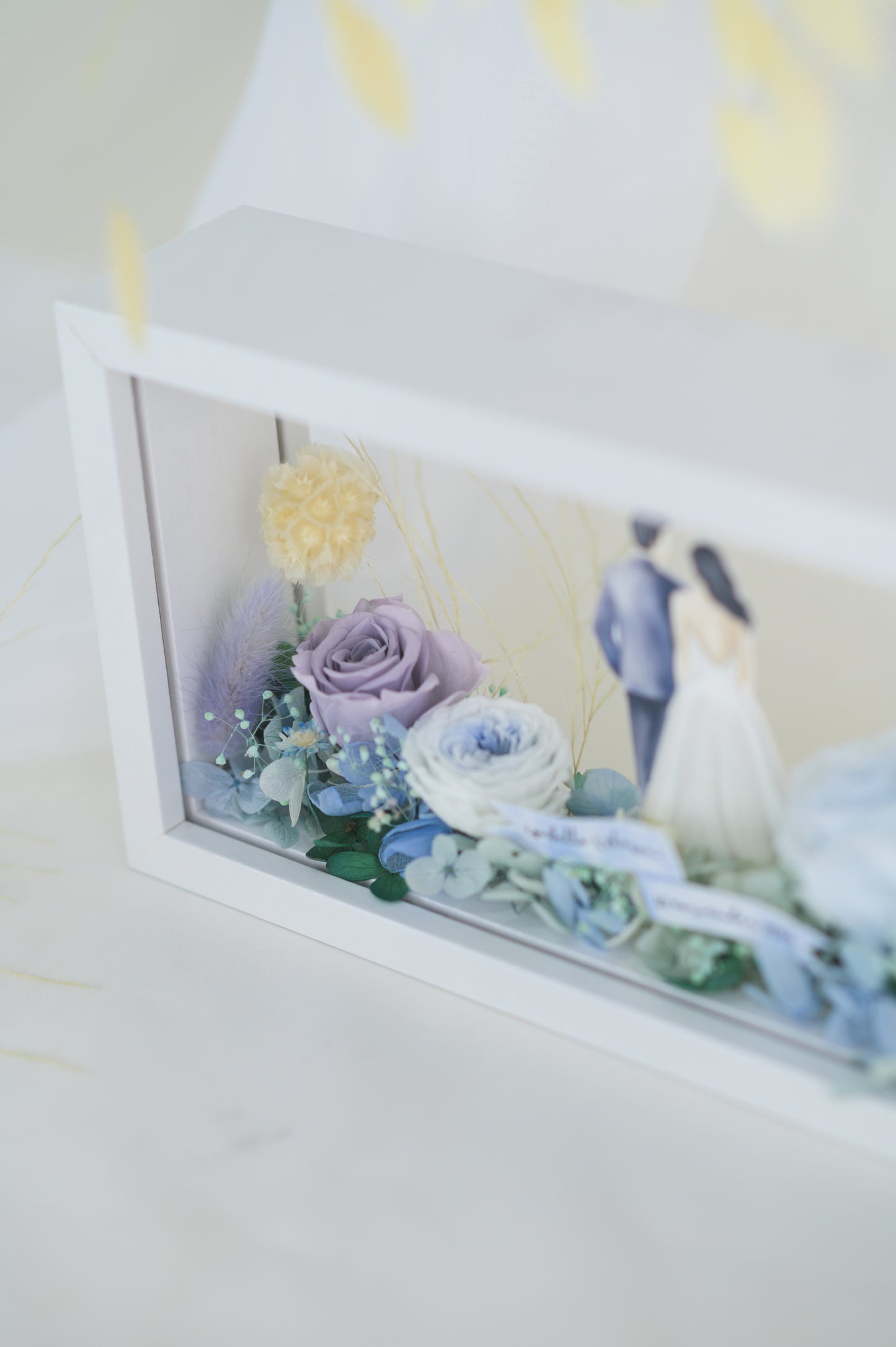A handcrafted wedding-themed frame featuring a bride and groom with preserved flowers in soft lavender, blue, and yellow tones, displayed in a double-sided glass frame for a 360-degree view