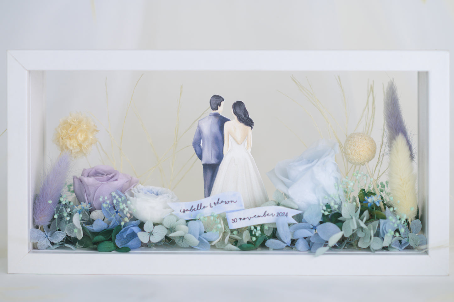 A handcrafted wedding-themed frame featuring a bride and groom with preserved flowers in soft lavender, blue, and yellow tones, displayed in a double-sided glass frame for a 360-degree view