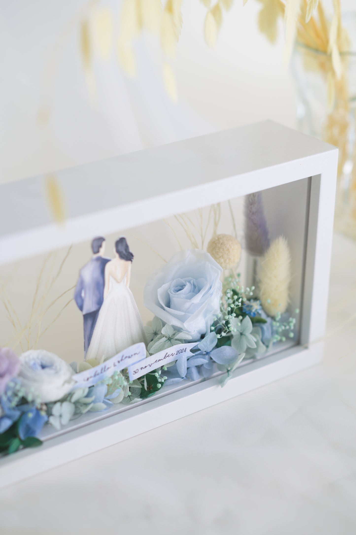 A handcrafted wedding-themed frame featuring a bride and groom with preserved flowers in soft lavender, blue, and yellow tones, displayed in a double-sided glass frame for a 360-degree view