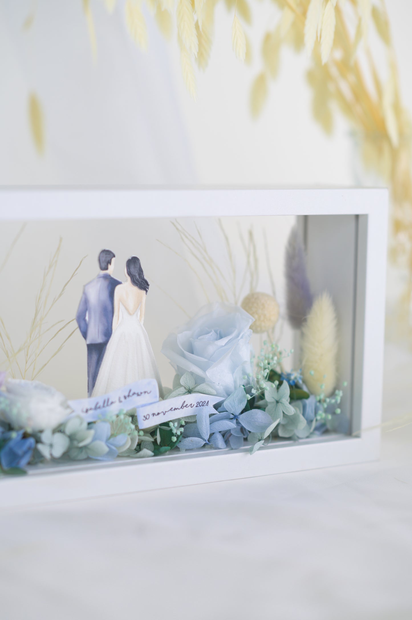 A handcrafted wedding-themed frame featuring a bride and groom with preserved flowers in soft lavender, blue, and yellow tones, displayed in a double-sided glass frame for a 360-degree view